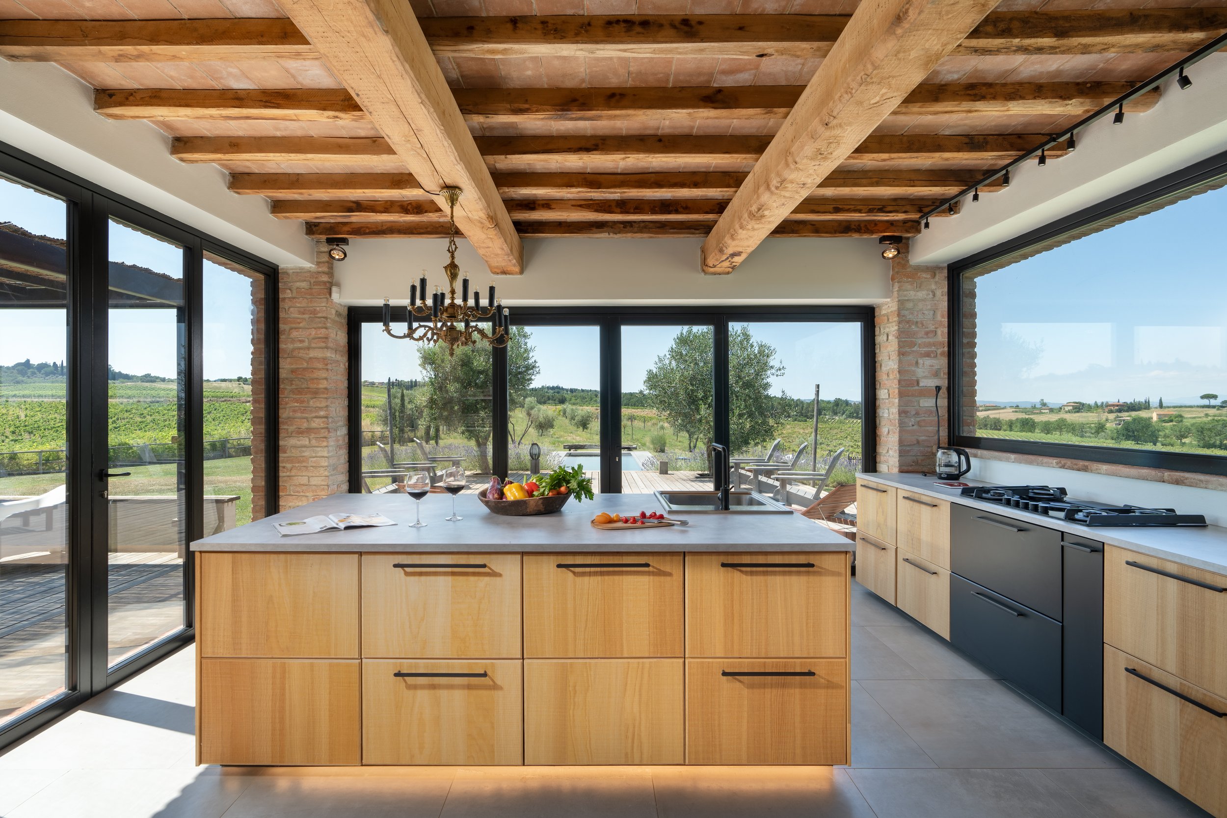 Modern kitchen with wooden cabinets, large windows showing outdoor view, kitchen island with fruit and wine glasses, dining area with chandelier, and outdoor patio.