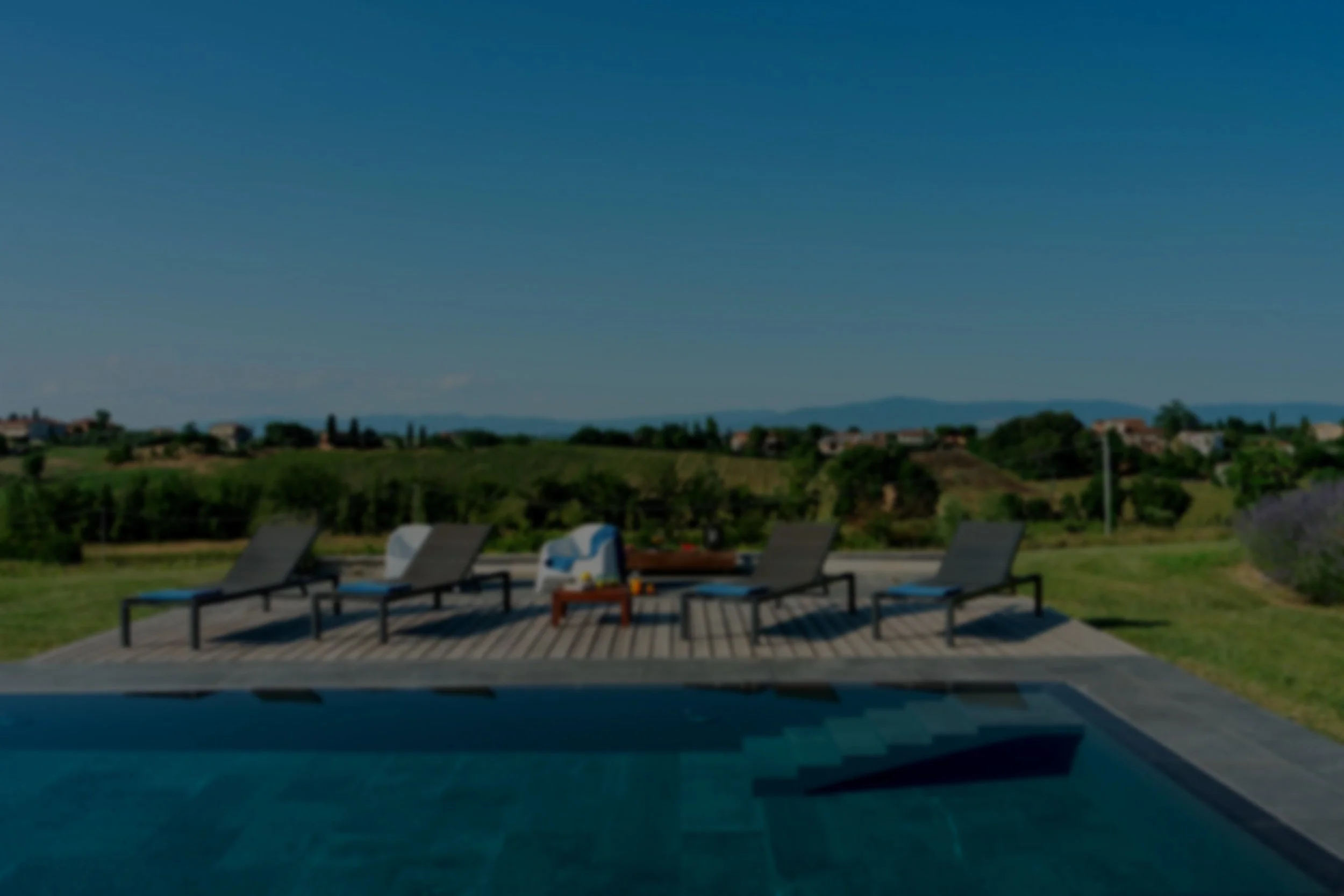 Outdoor pool area with four lounge chairs on a wooden deck, and a view of green rolling hills under a clear blue sky.