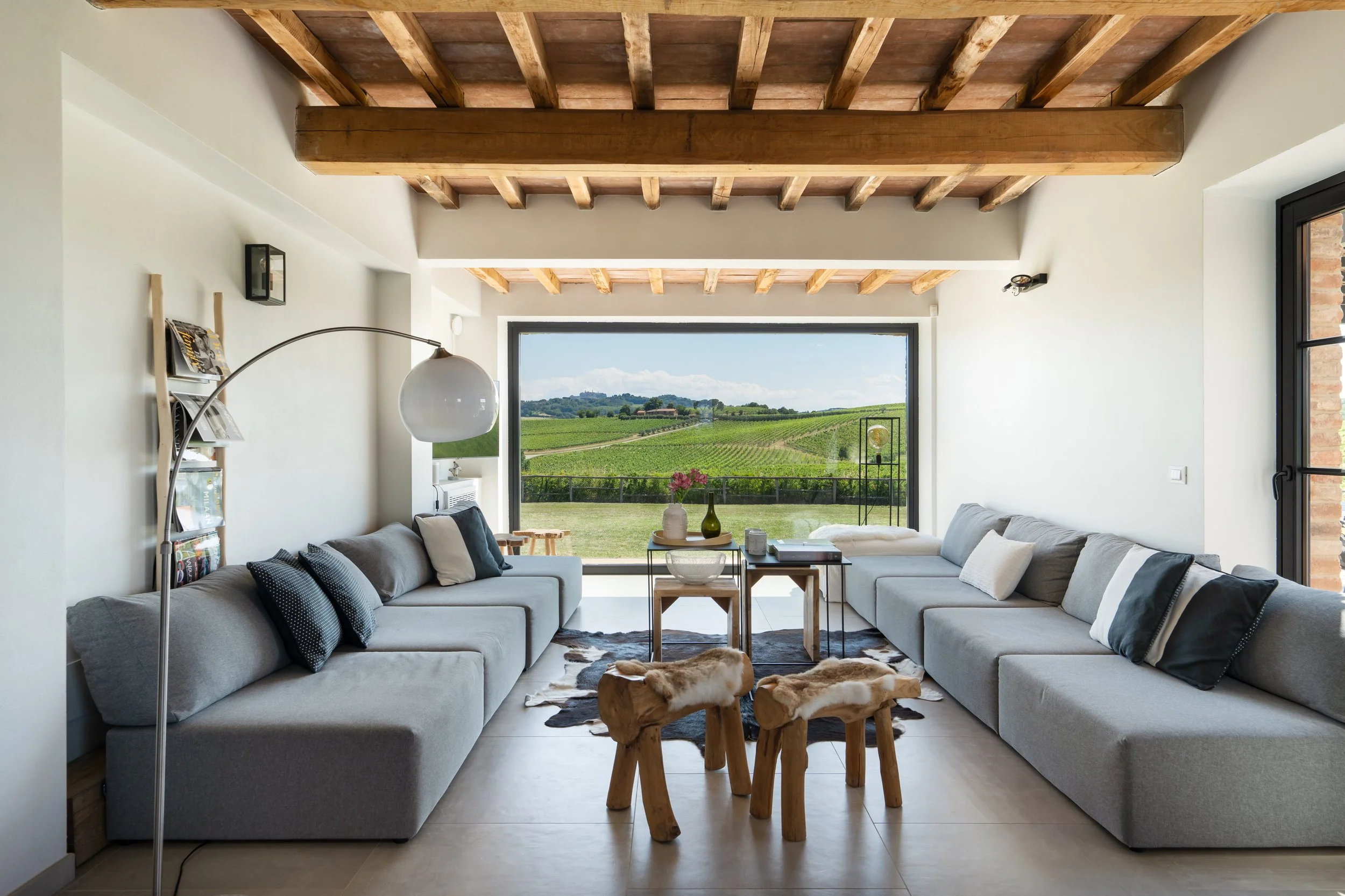 Modern living room with large windows showing a scenic view of green vineyards and hills, featuring two long gray sofas, a cowhide rug, small wood stools, and minimalistic decor.