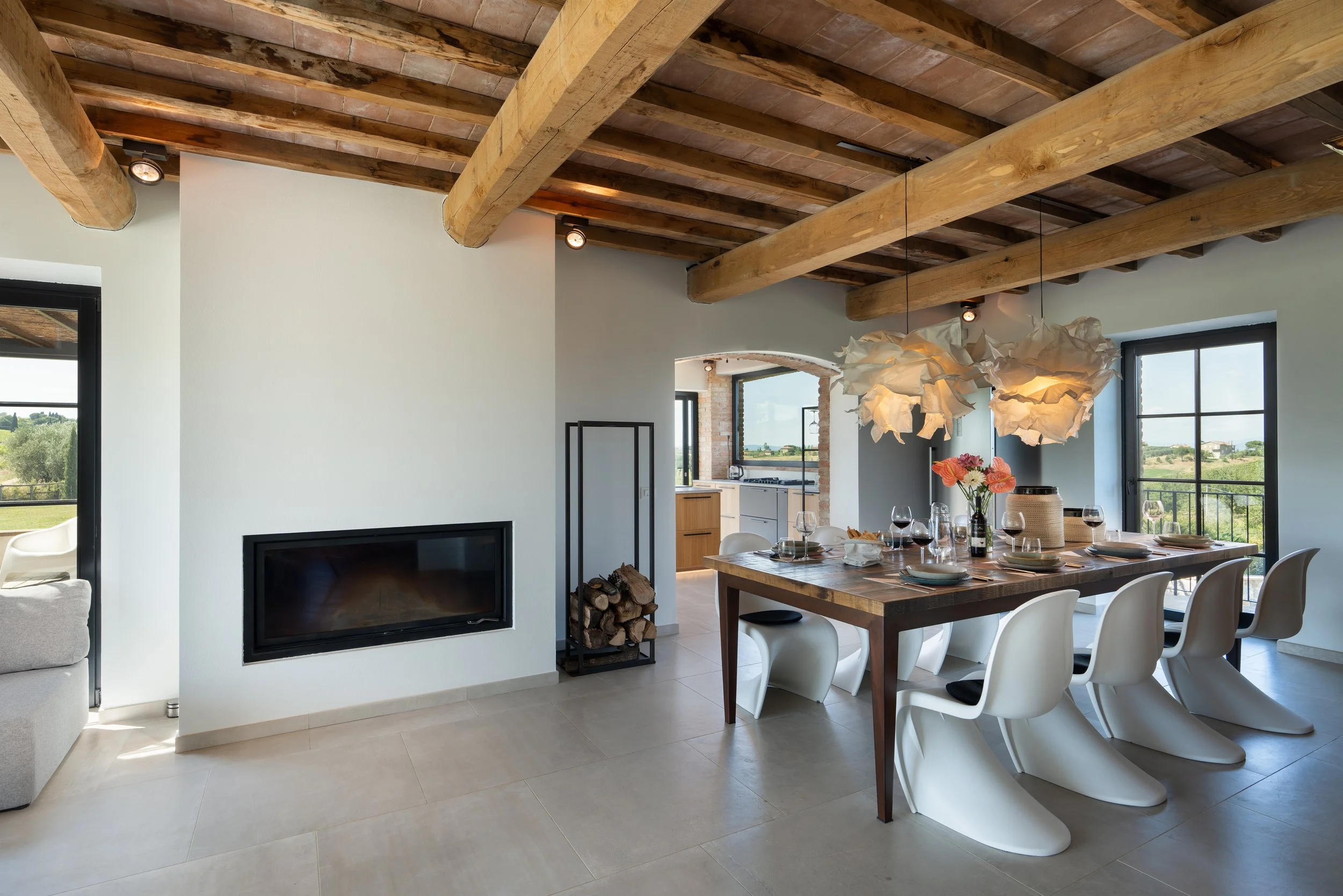 A dining room with a large wooden table, white chairs, and modern light fixtures. There is a view of a kitchen through an arched opening, and a large window with black frames showing a scenic outdoor landscape.