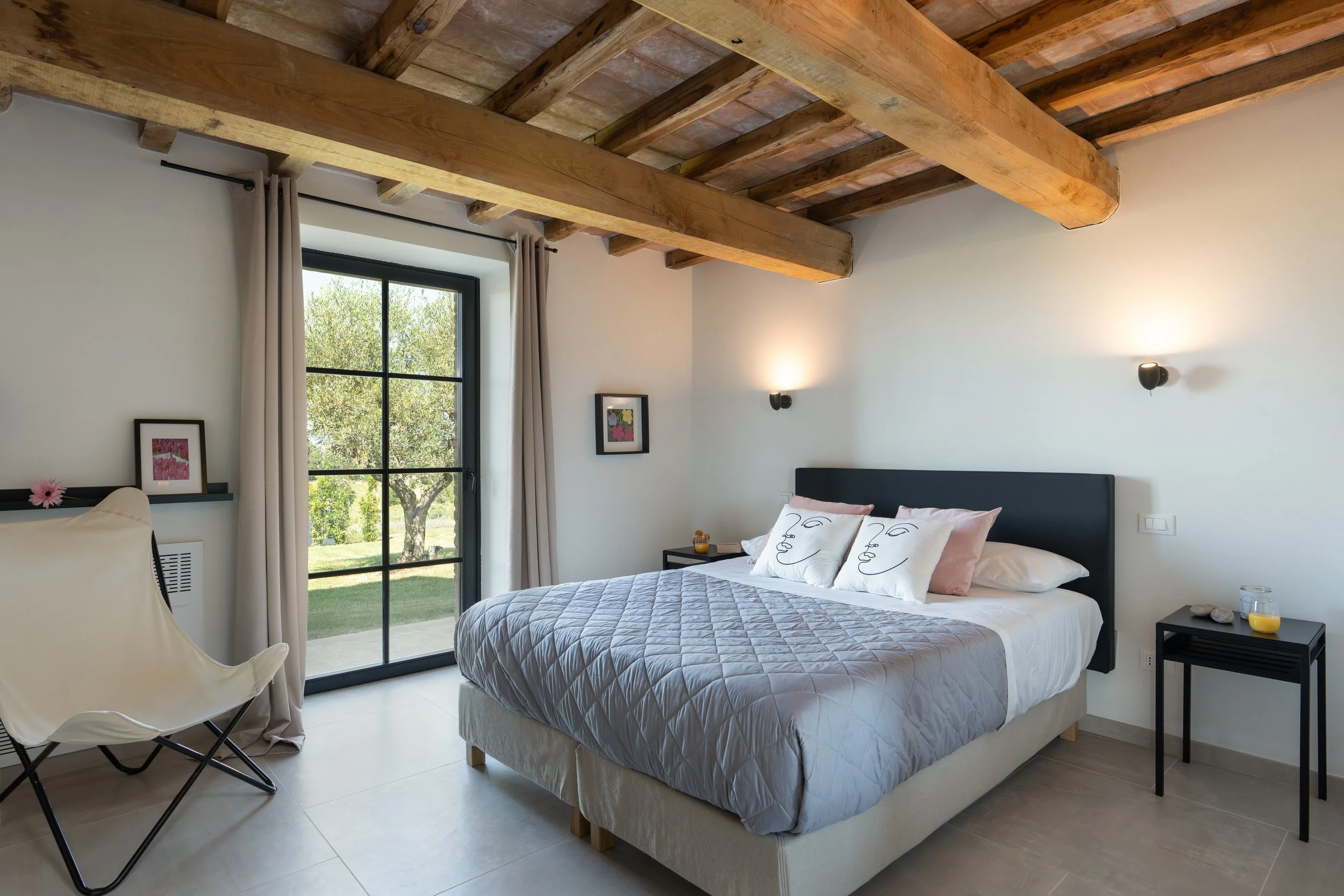 A bedroom with a large bed, white bedding, and decorative pillows featuring abstract line art faces. There are black nightstands on each side of the bed, with one holding a glass of orange juice. The room has a glass door leading outside, with beige 