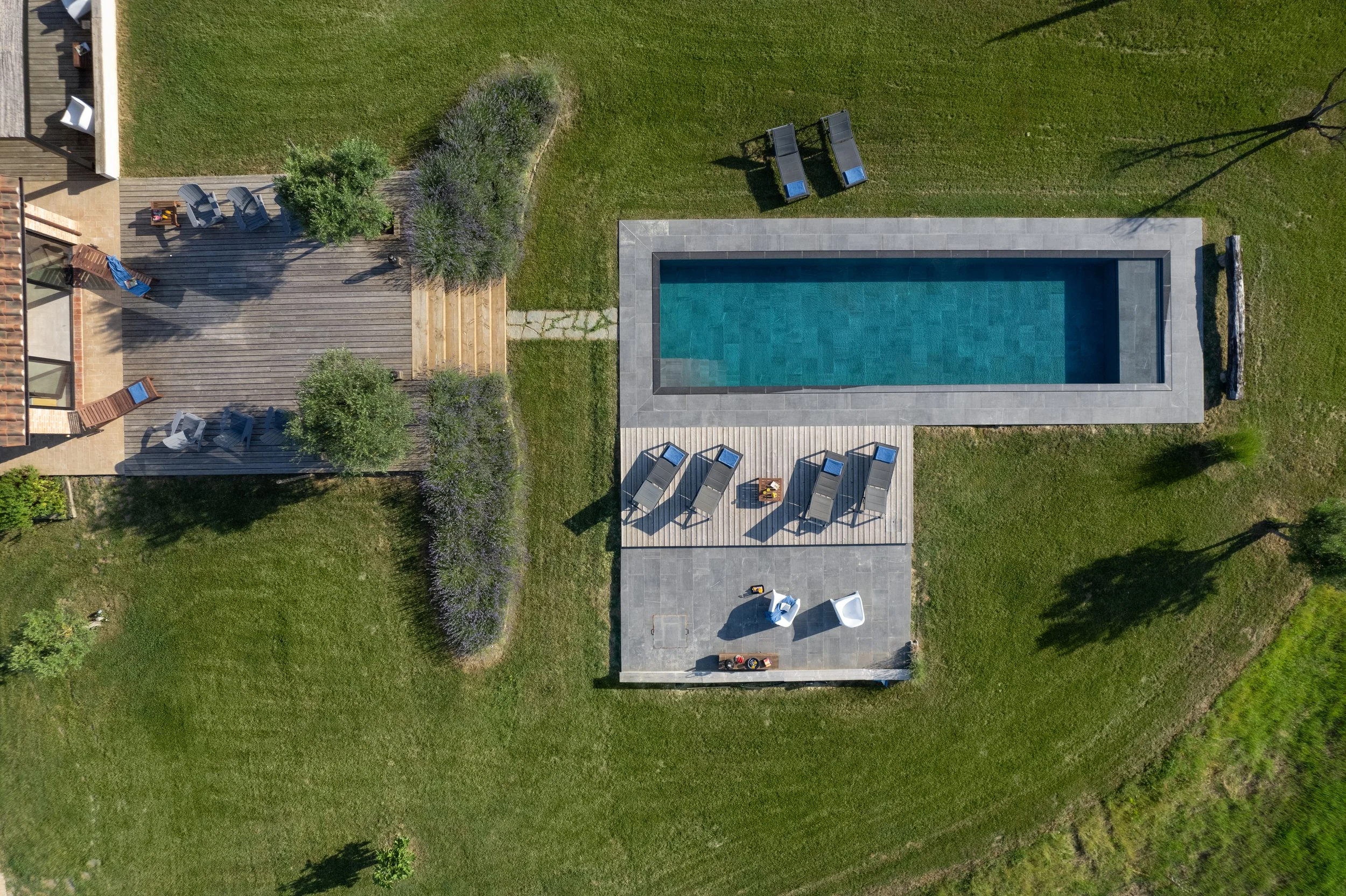 Aerial view of a backyard featuring a rectangular swimming pool, wooden deck with lounge chairs, a grassy lawn with landscaping, and a paved patio area with outdoor furniture.