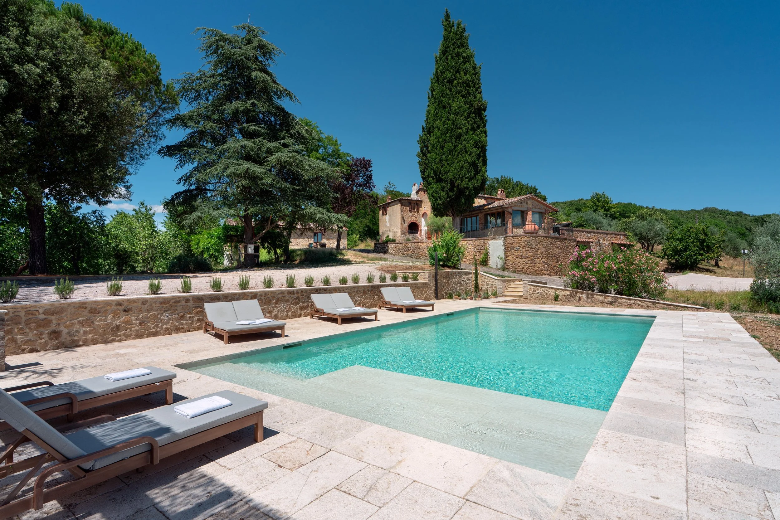 A serene outdoor swimming pool surrounded by lounge chairs with towels, stone patio, lush green trees, bushes, and rustic houses under a bright blue sky.