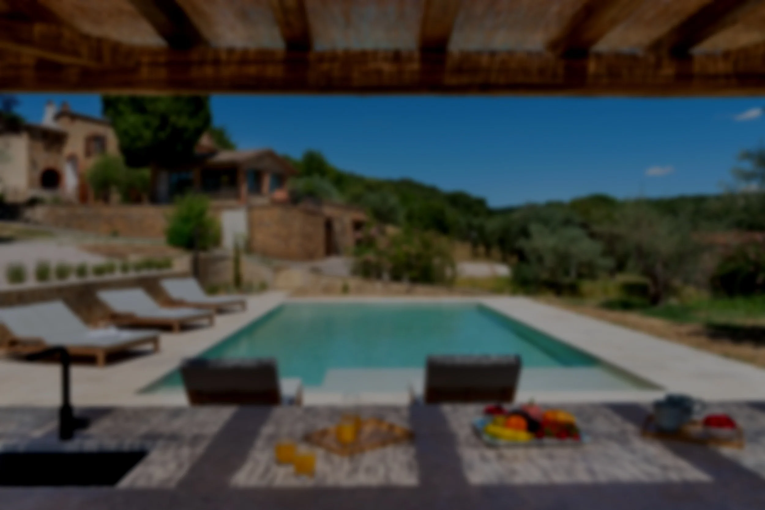 Outdoor swimming pool with lounge chairs, a patio table with food and drinks, and a scenic hillside village in the background under a clear blue sky.