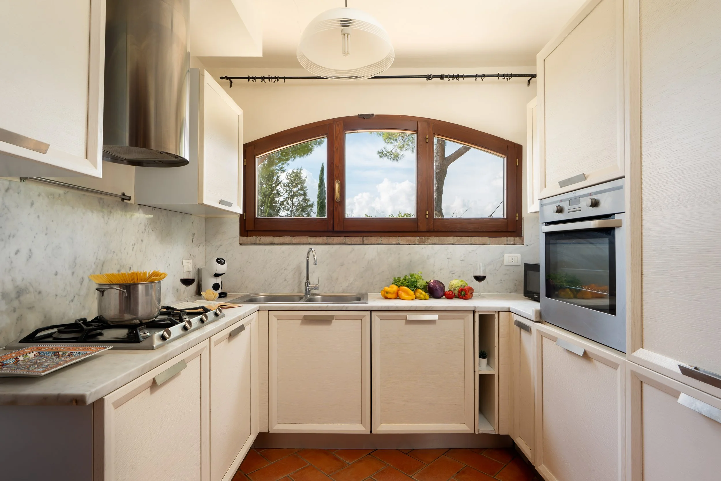 Kitchen with a marble countertop, white cabinets, a window with a wooden frame, stove with a pot, wine glasses, vegetables and herbs, a microwave, and a built-in oven.