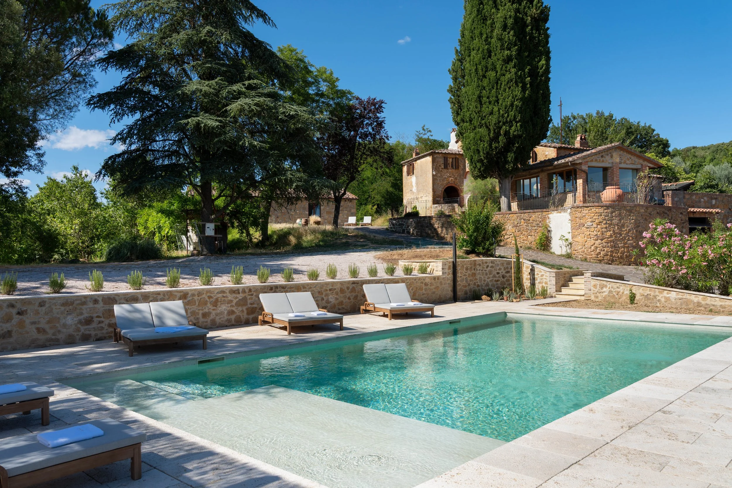 A swimming pool with white lounge chairs on a stone deck, surrounded by lush green trees and rustic stone buildings under a clear blue sky.