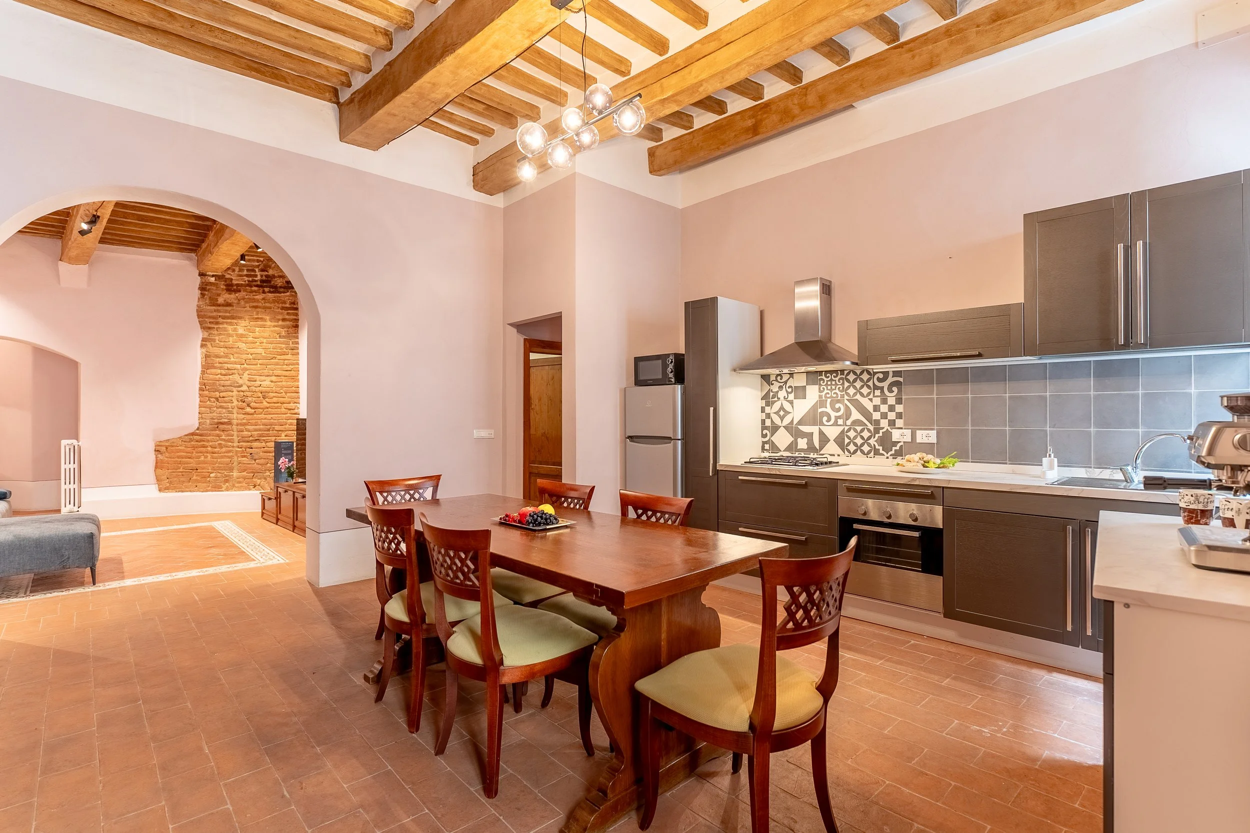 Kitchen and dining area with wooden ceiling beams, a wooden dining table with six chairs, a built-in kitchen with dark gray cabinets, stainless steel appliances, a patterned tile backsplash, and a partial view of a living room with brick archway and 
