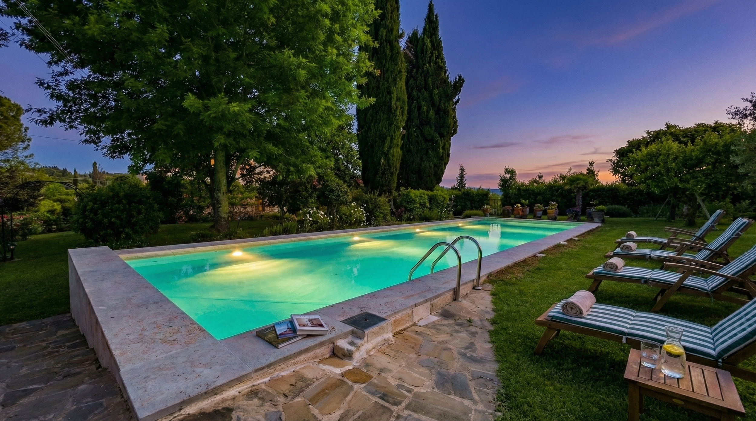 An outdoor swimming pool illuminated at dusk, surrounded by lounge chairs with rolled towels, glass pitcher with water and lemon, and lush trees and bushes in the background.