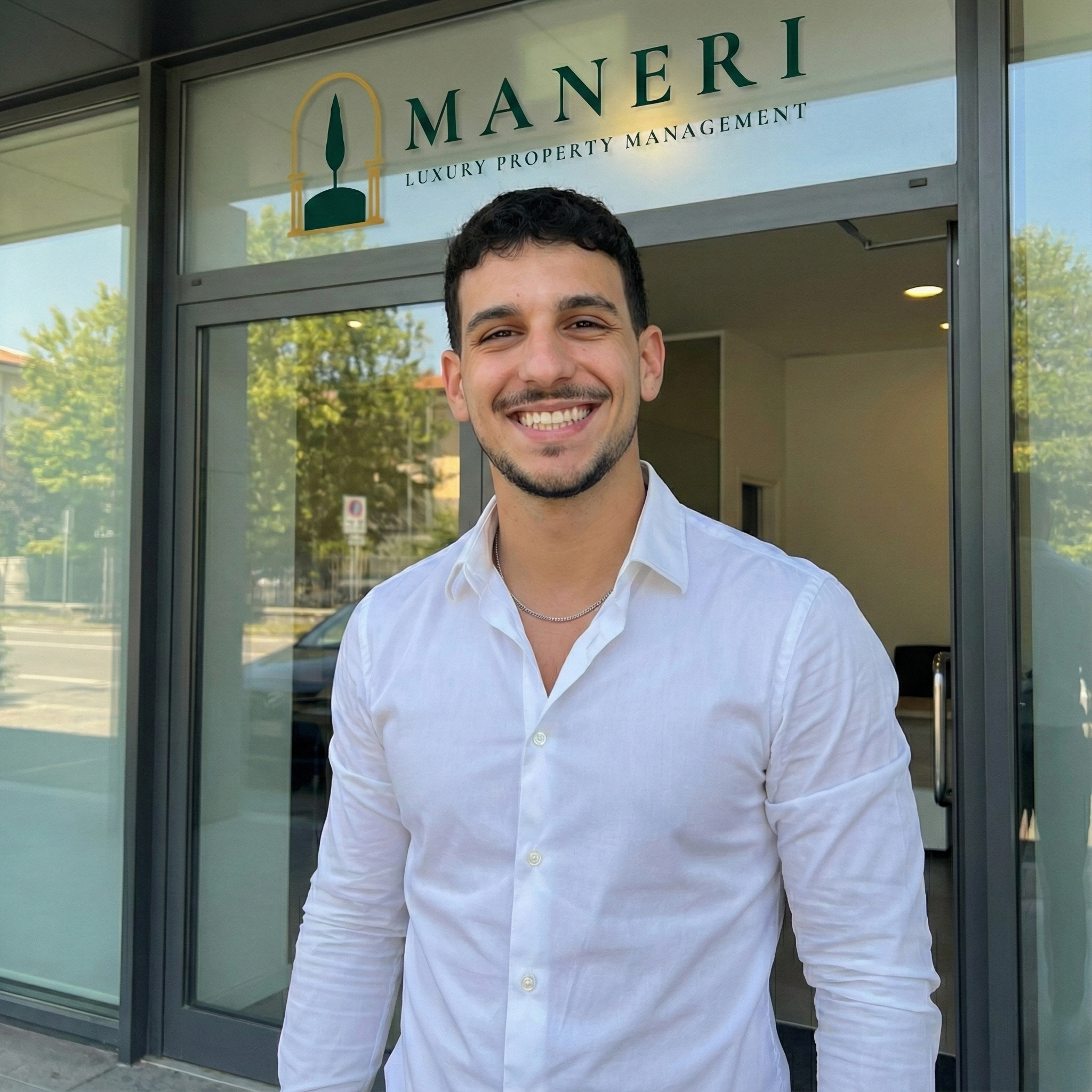 A smiling young man in a white button-up shirt standing outside a building with a sign that reads 'MANERI LUXURY PROPERTY MANAGEMENT'. The building has large glass windows reflecting trees and parked cars.