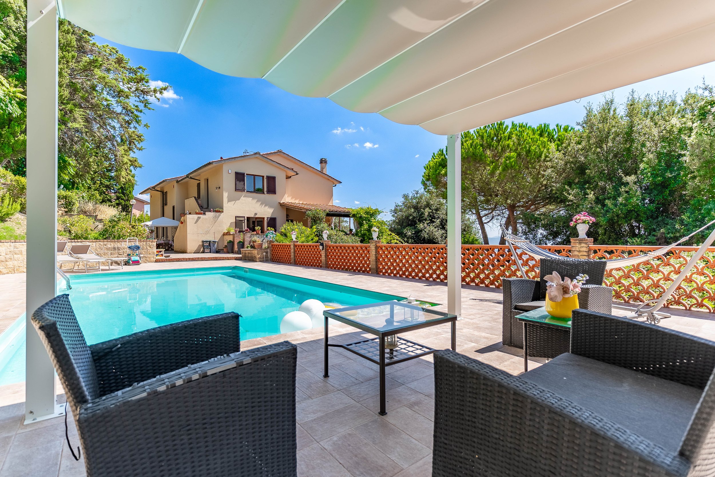 A backyard with a swimming pool, outdoor seating with black wicker chairs, and a house in the background under a sunny blue sky.