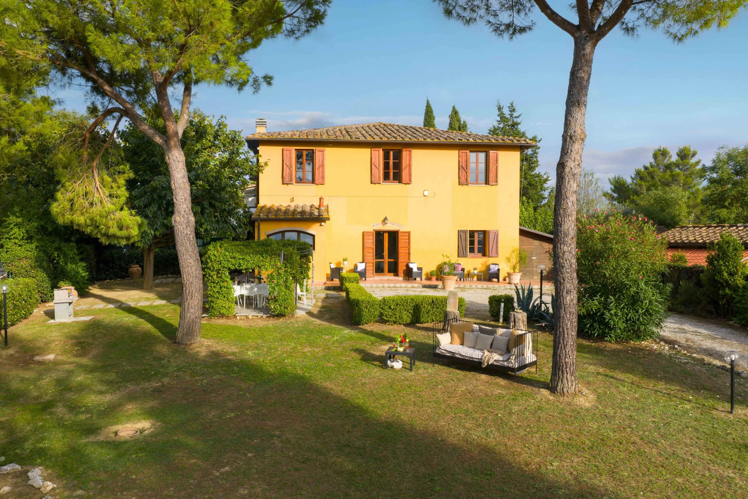 A yellow two-story house with wooden shutters, surrounded by trees, bushes, and outdoor seating areas, on a sunny day.