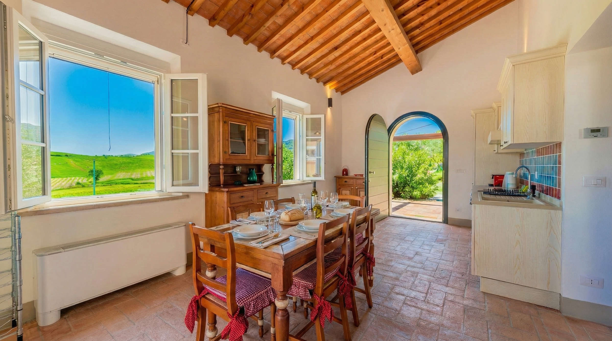 Sunlit rustic kitchen and dining area with wooden ceiling and open windows, set for a meal with plates, glasses, and bread on a wooden table.