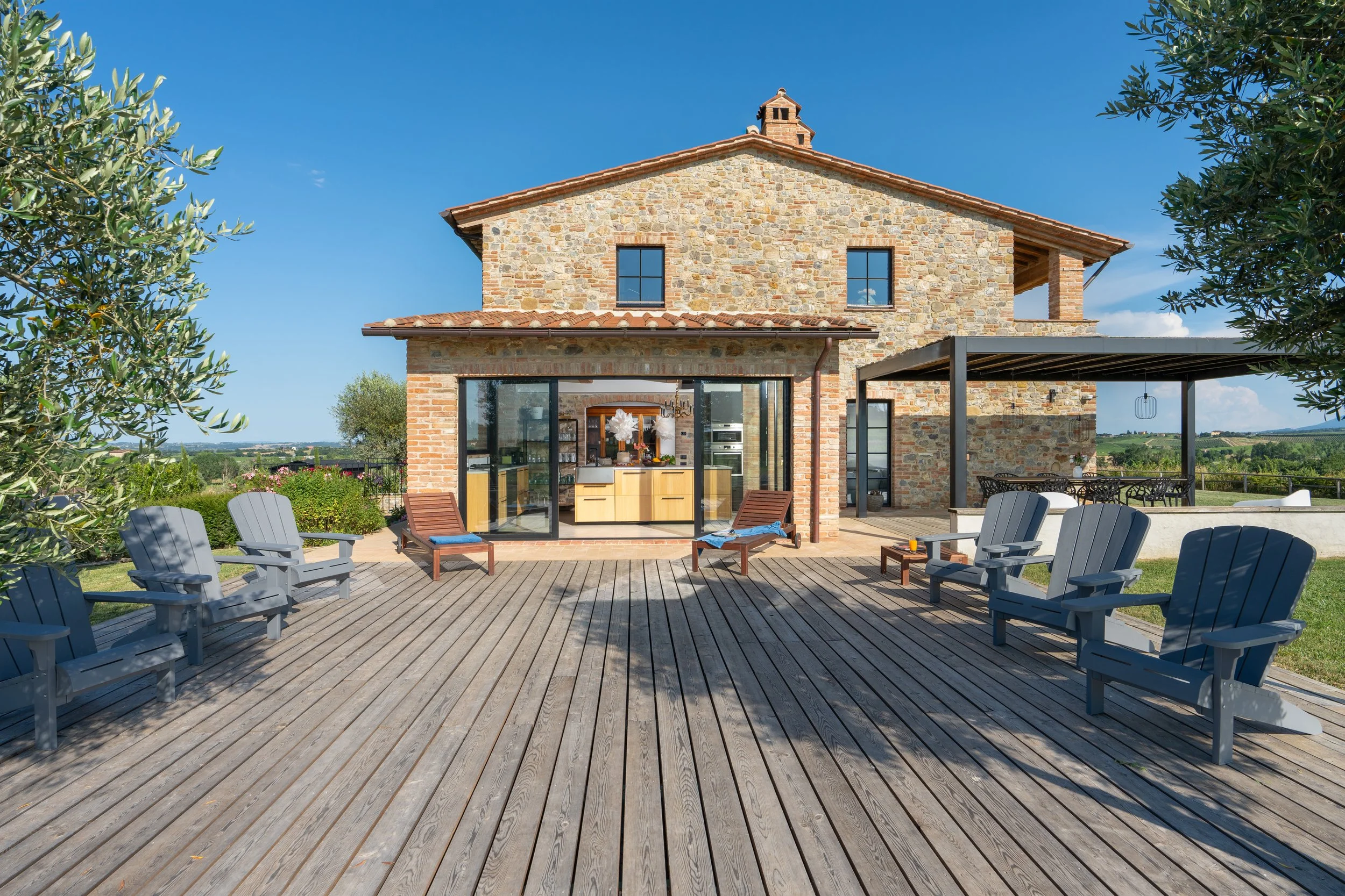 A rustic stone house with a wooden deck outdoor patio, featuring several gray and wooden chairs facing the house, overlooking a scenic countryside landscape under a clear blue sky.