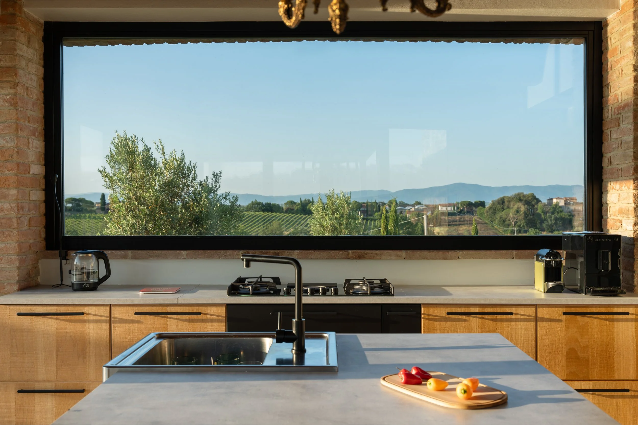 Modern kitchen with a large window overlooking a scenic landscape with trees and mountains, countertop with a sink, gas stove, coffee maker, kettle, and a cutting board with vegetables.