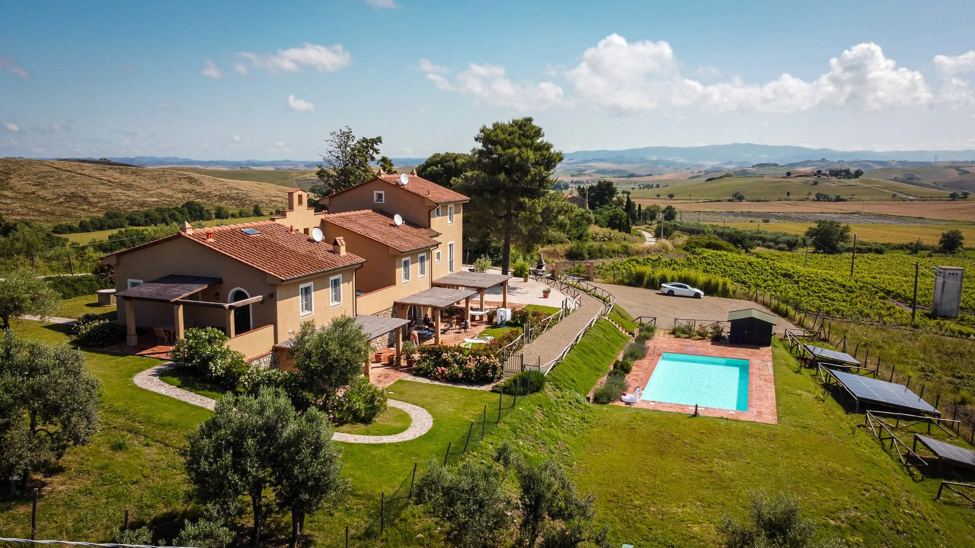 A scenic countryside estate with beige houses, a swimming pool, patio area, and solar panels, surrounded by lush green gardens and rolling hills under a blue sky with scattered clouds.