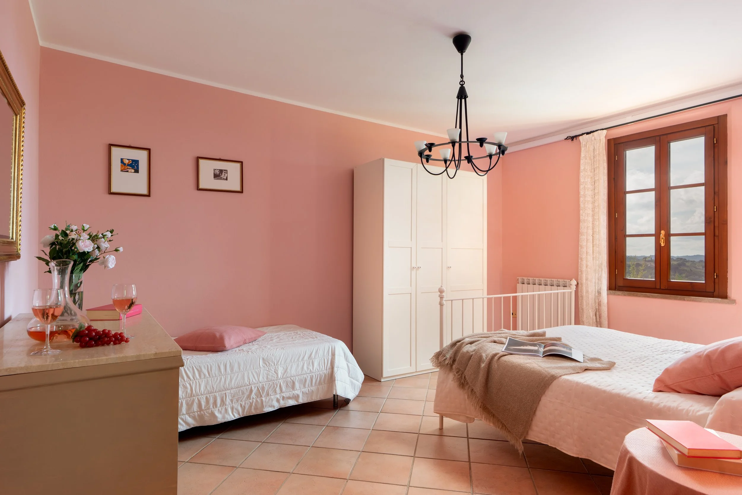 Bedroom with pink walls, two beds, a white wardrobe, a window with a view, a chandelier, and decorative items including books, glasses, and flowers.
