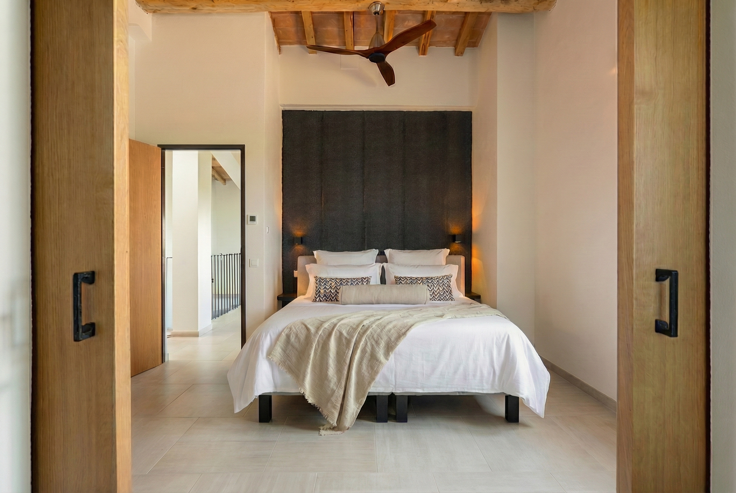Modern bedroom with white bedding, black headboard, decorative pillows, beige blanket, and wooden ceiling beams, viewed through sliding wooden doors.