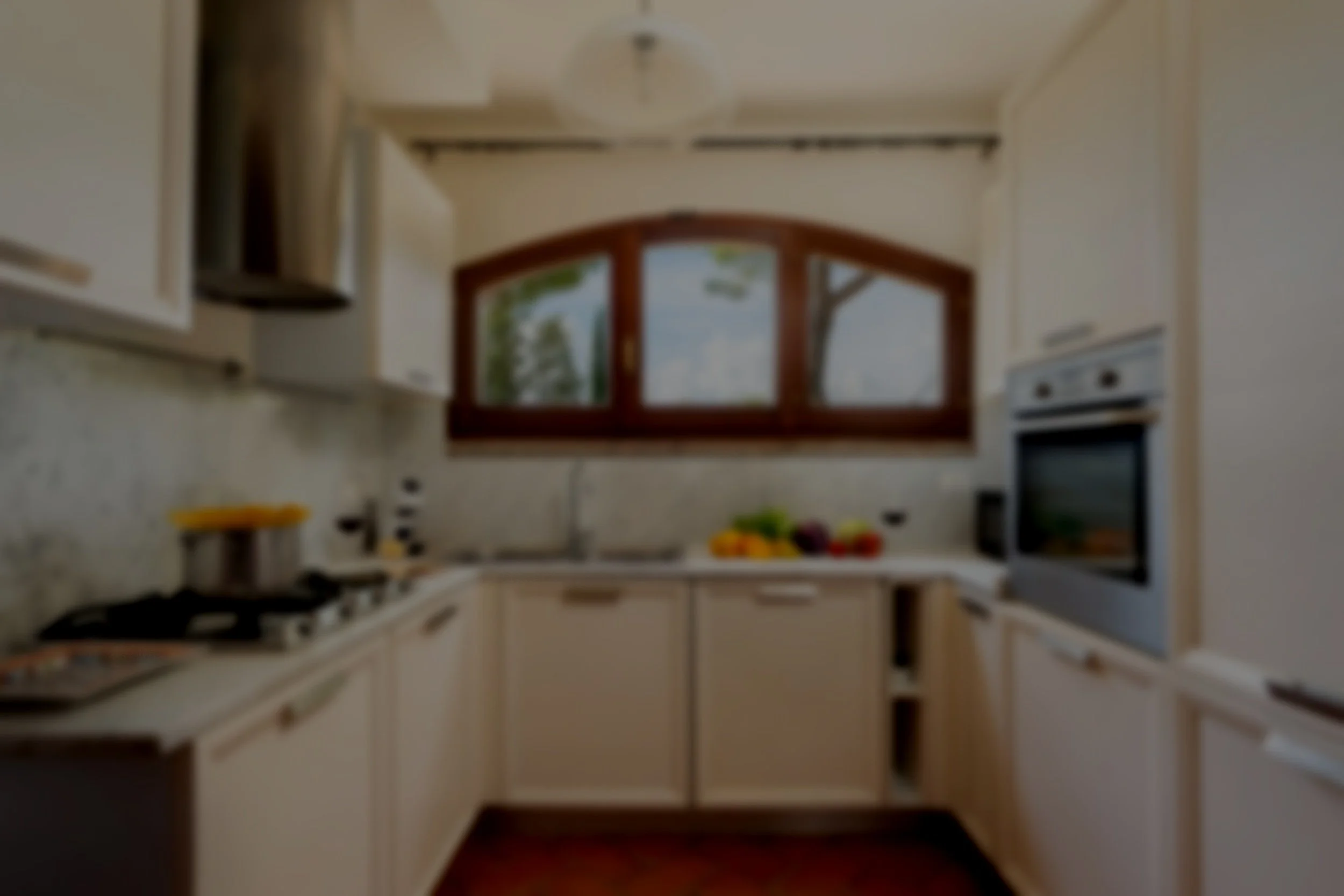 Blurry kitchen with cream-colored cabinets, windows showing trees outside, and a variety of kitchen appliances and food on the countertops.