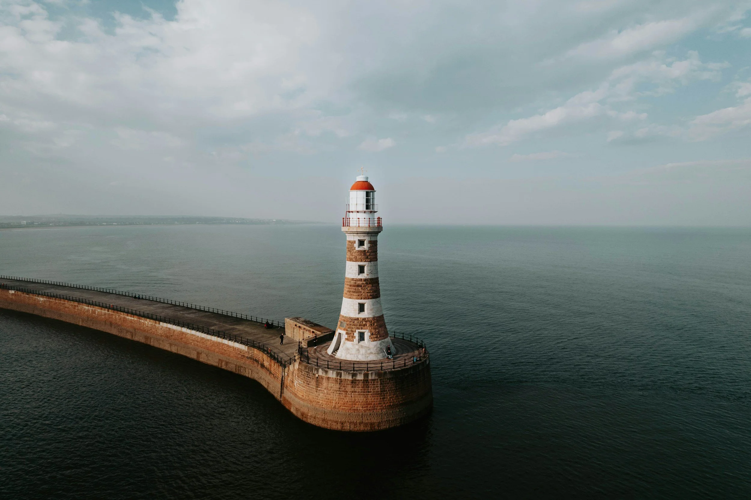 A lighthouse situated at the end of a curved stone pier extending into calm water, with a partly cloudy sky overhead.
