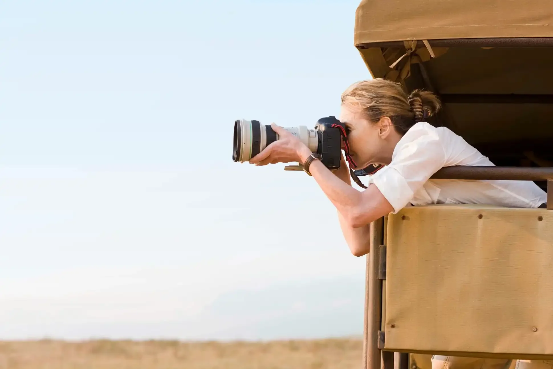 Woman with blonde hair in a ponytail taking a photograph from a wooden lookout with a camera fitted with a large telephoto lens, looking outward from the side of the structure at the outdoors with a clear sky and distant field.