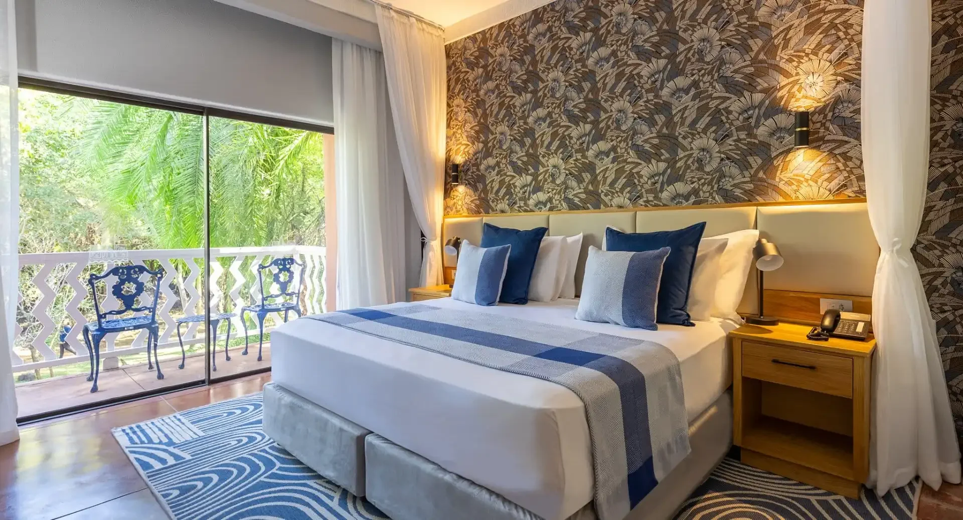 A cozy hotel bedroom with a large bed, decorated with multiple blue and white pillows and a striped bedspread. The bed is beside a wooden nightstand holding a telephone and a lamp. The room has a patterned wallpaper behind the bed, white curtains, and a large sliding glass door leading to a balcony with outdoor chairs and lush green trees outside.