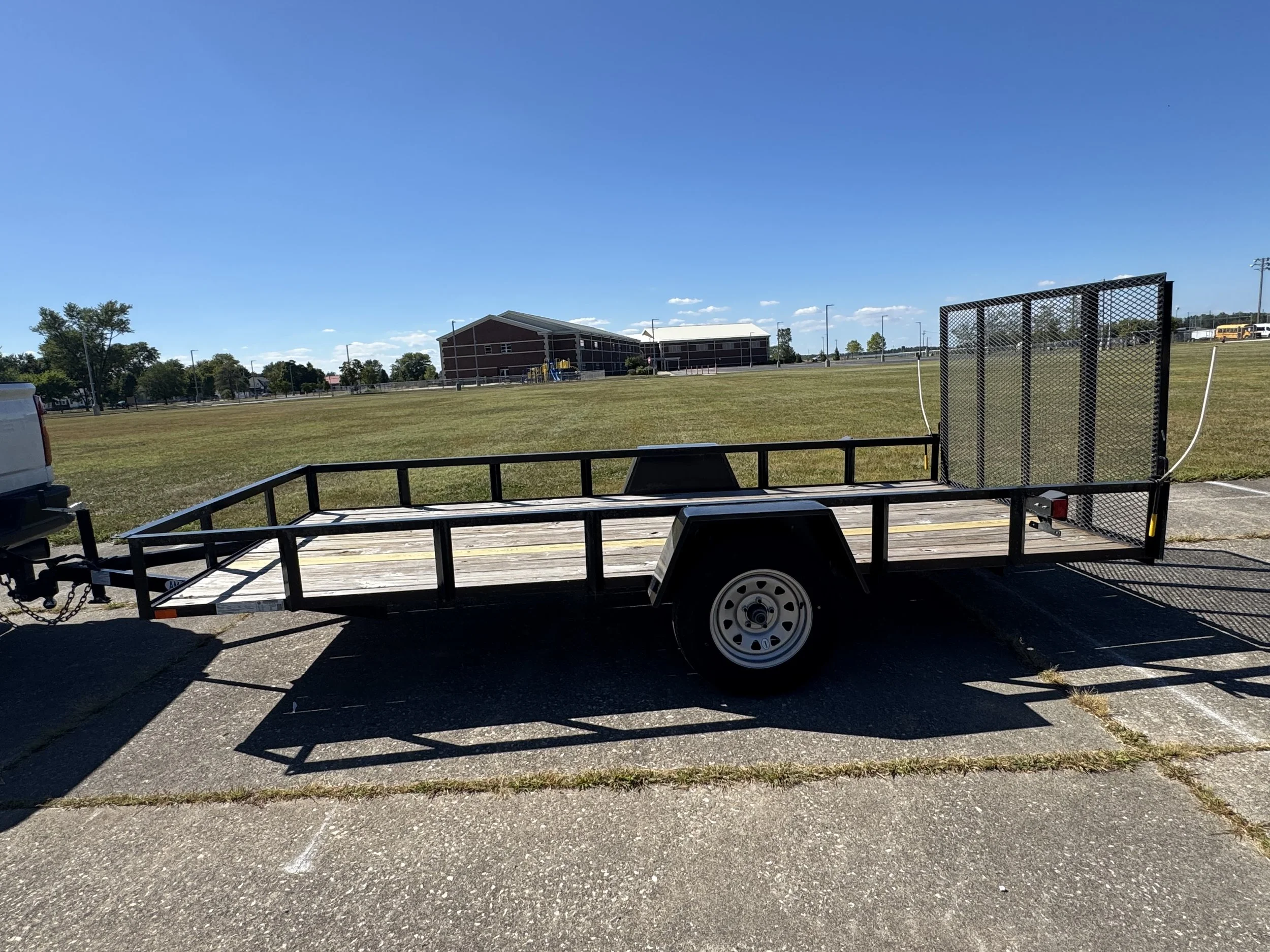 Empty flatbed trailer parked on a concrete surface in an open grassy area with a school building and clear blue sky in the background.