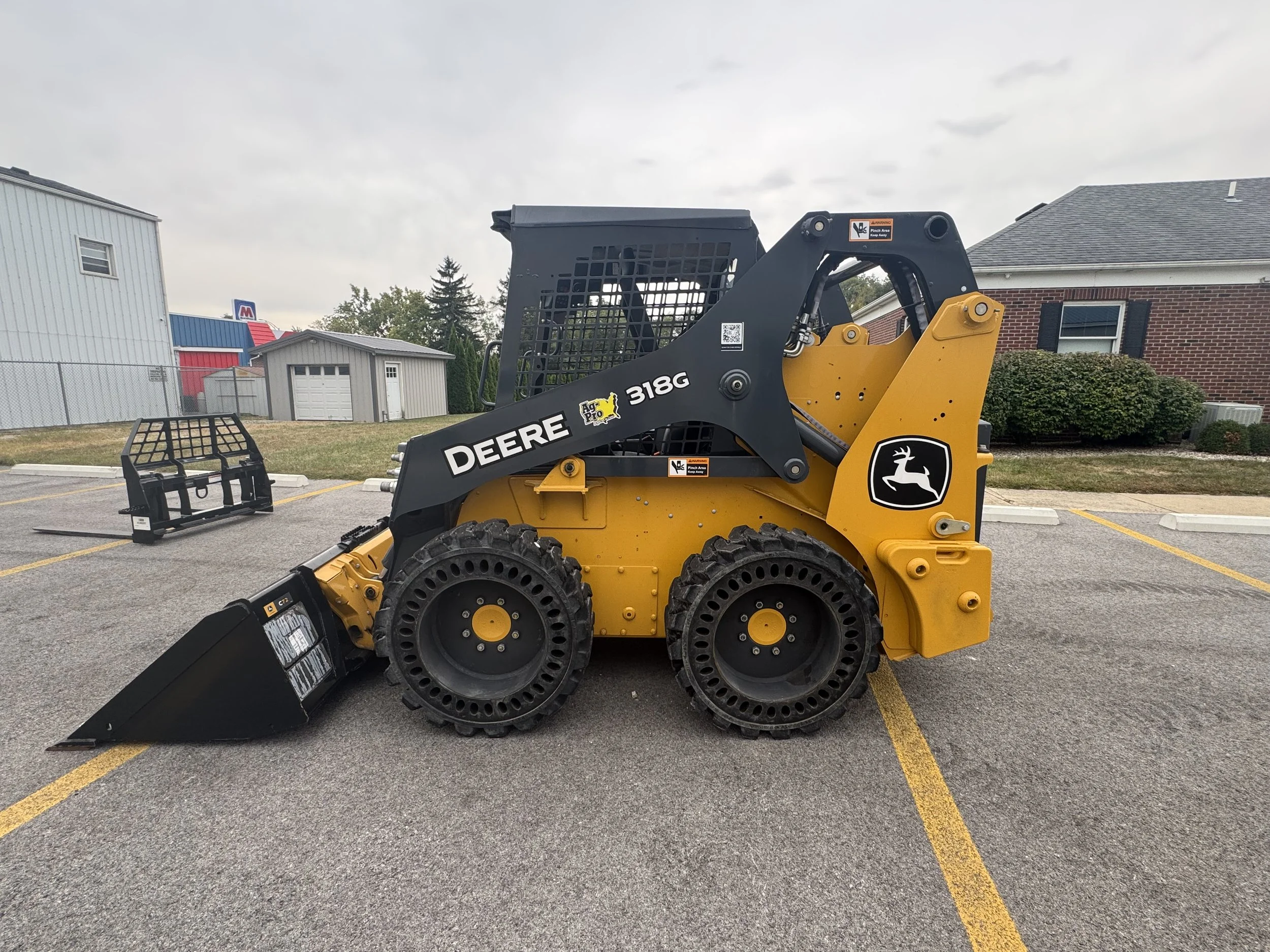 A John Deere skid steer loader in a parking lot with a wheel attachment nearby, and a building, bushes, and a cloudy sky in the background.