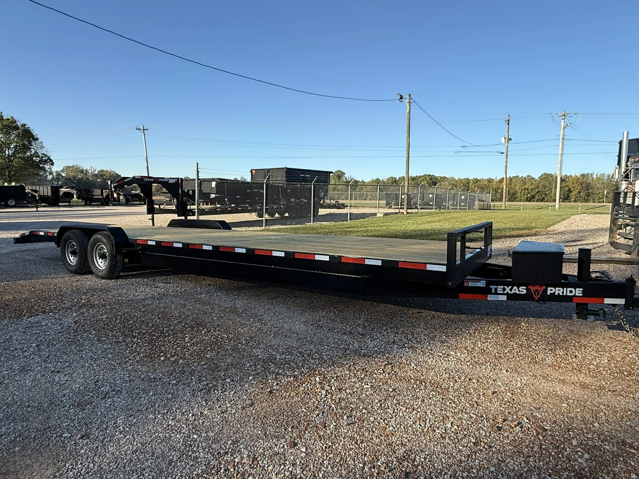 A black flatbed trailer with dual wheels, parked on a gravel lot with fencing and utility poles in the background.