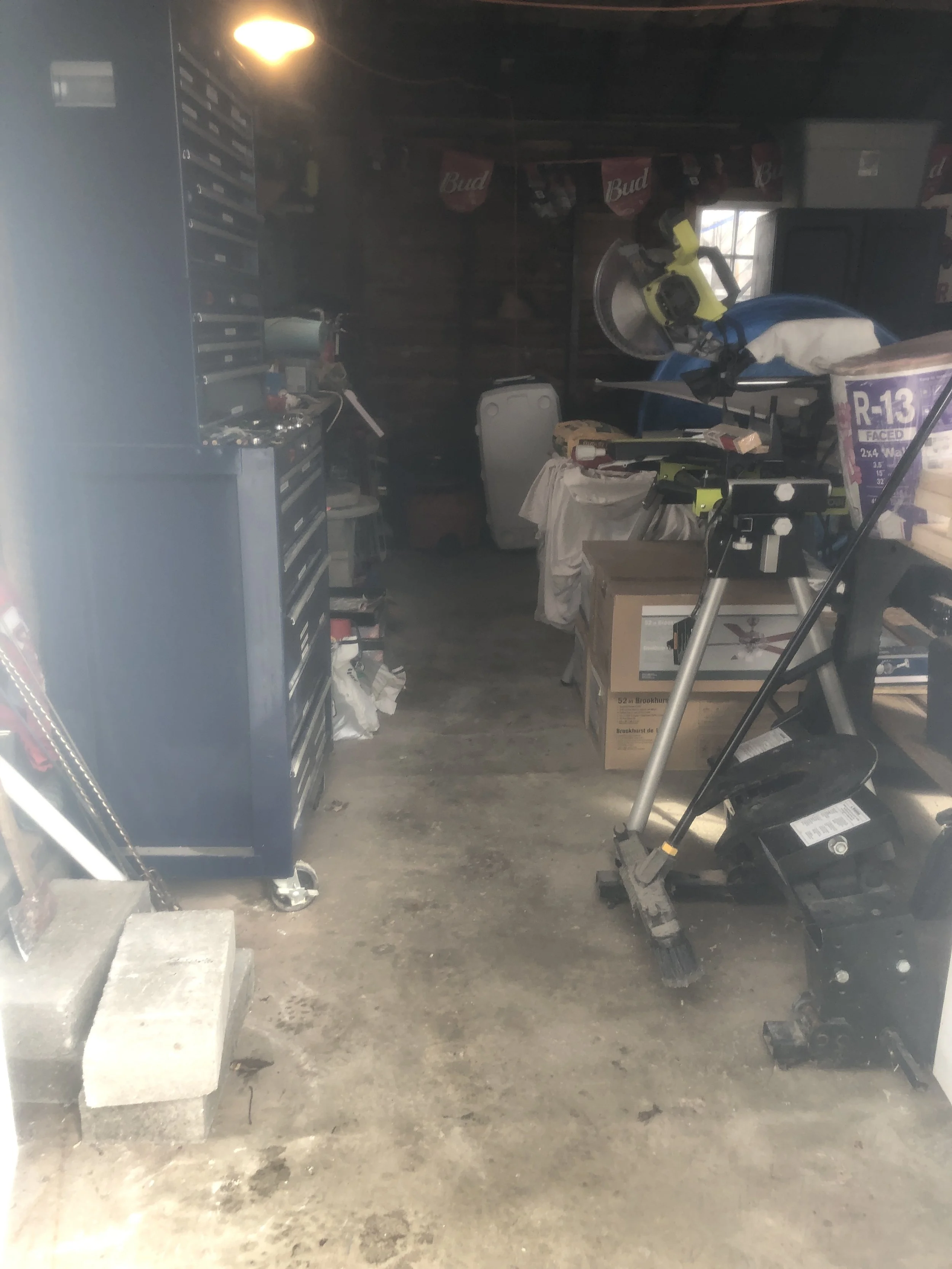 Open garage filled with various tools, equipment, and storage items, including a propane tank, yellow step ladder, metal trash can, and a cement mixer.