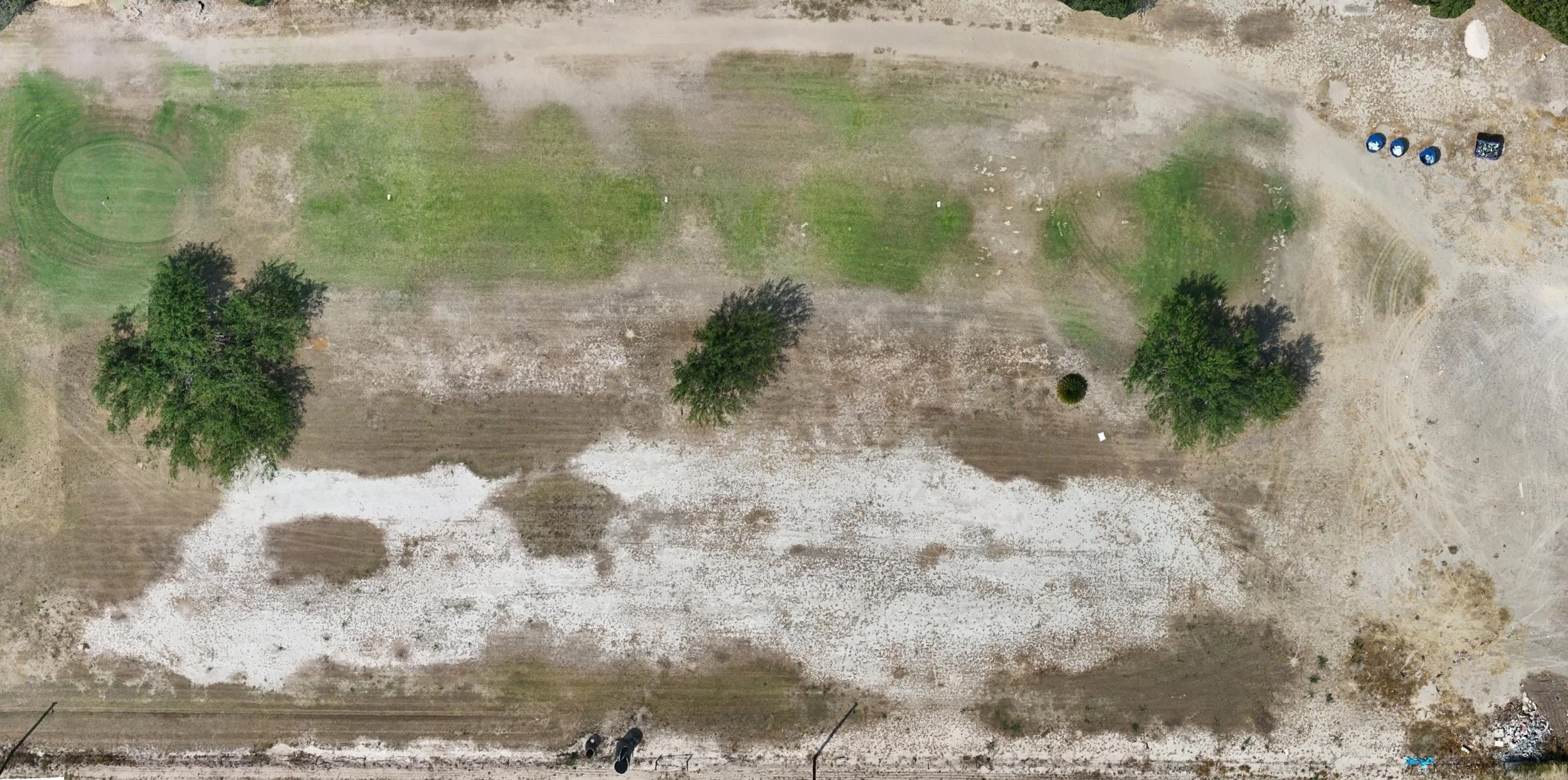 An aerial view of a field with patches of grass, trees, and exposed soil, along with some scattered equipment and structures in the background.