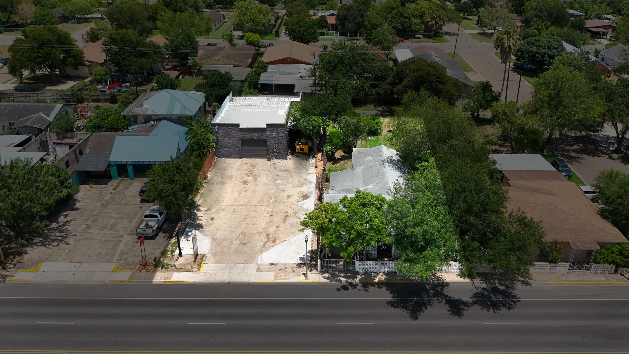 An aerial view of a residential neighborhood with houses surrounded by trees, a large empty parking lot in the foreground, and a commercial building with a garage and construction equipment in the center.