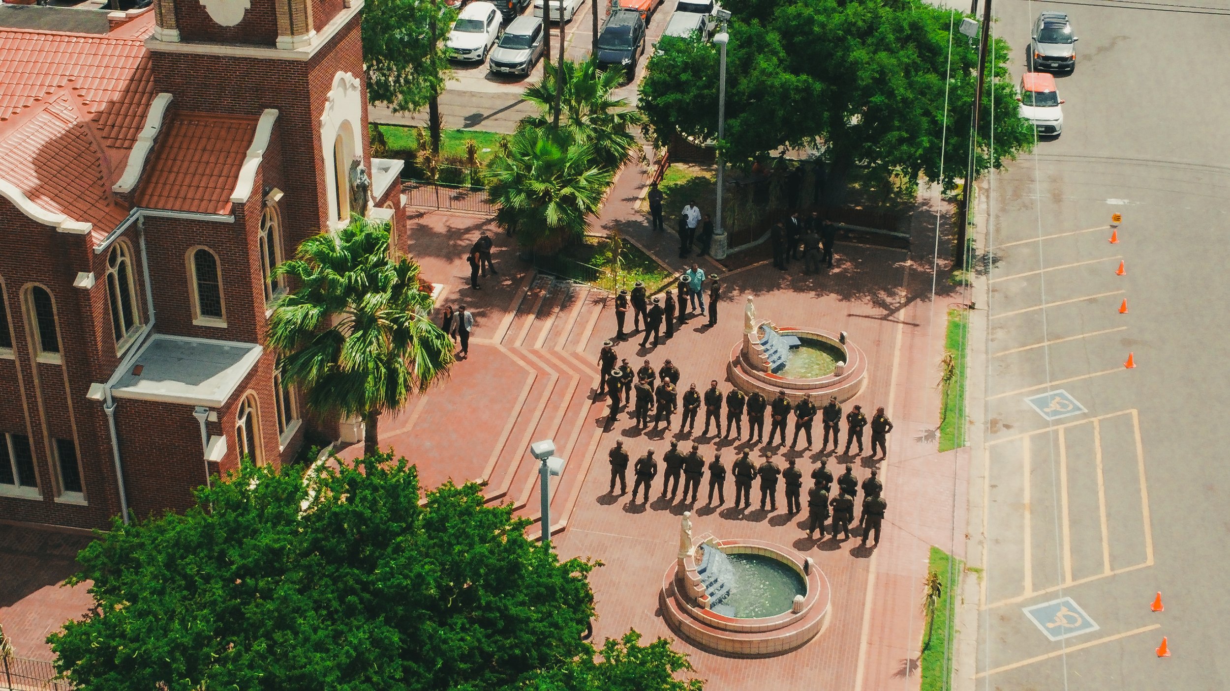 Aerial view of a group of uniformed officers standing in formation outside a brick church with a red tile roof, near a fountain with water spouting, trees, and parked cars.