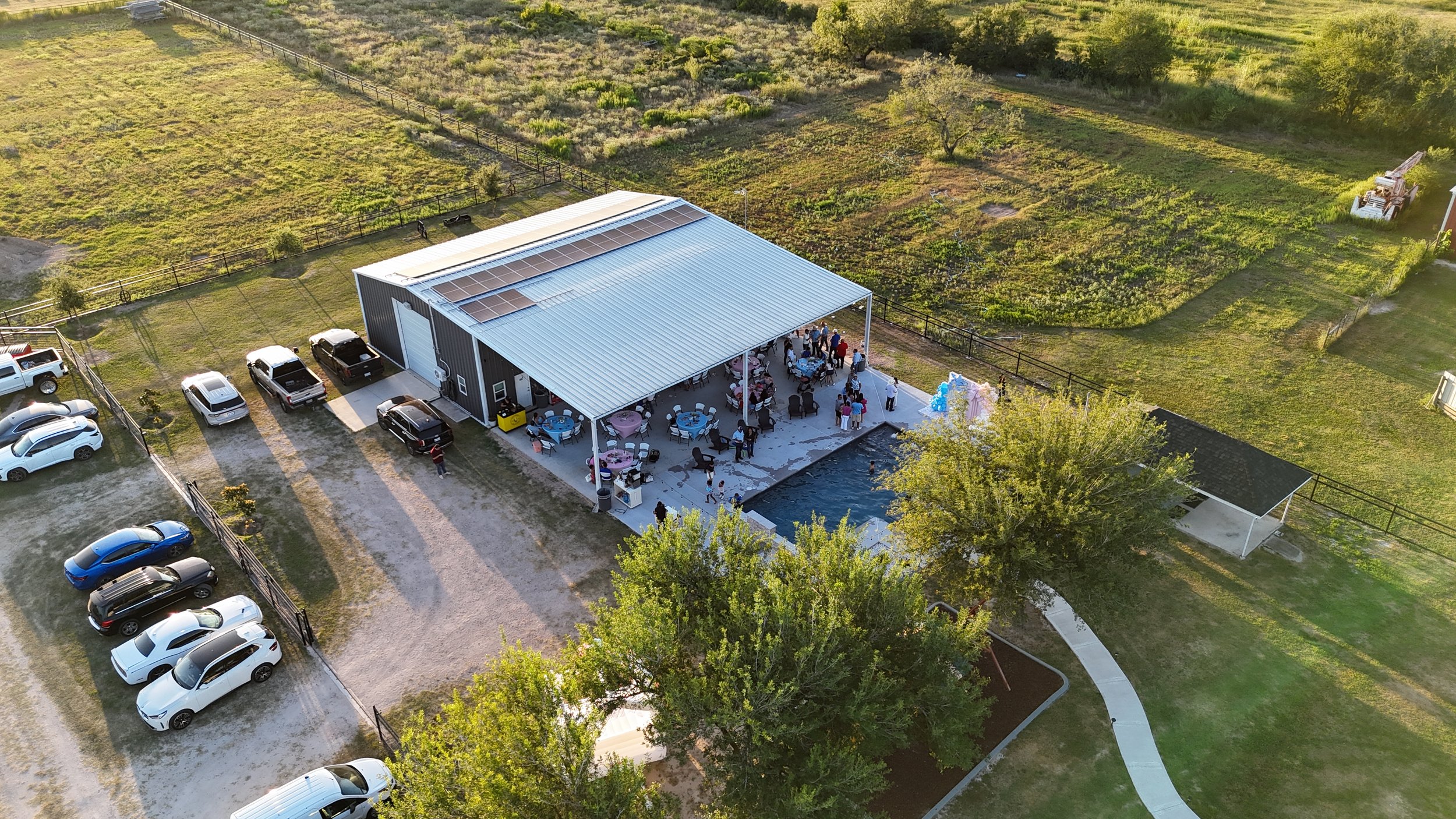 Aerial view of a backyard party with a metal building, outdoor tables, and a small pool, surrounded by greenery and parked cars.