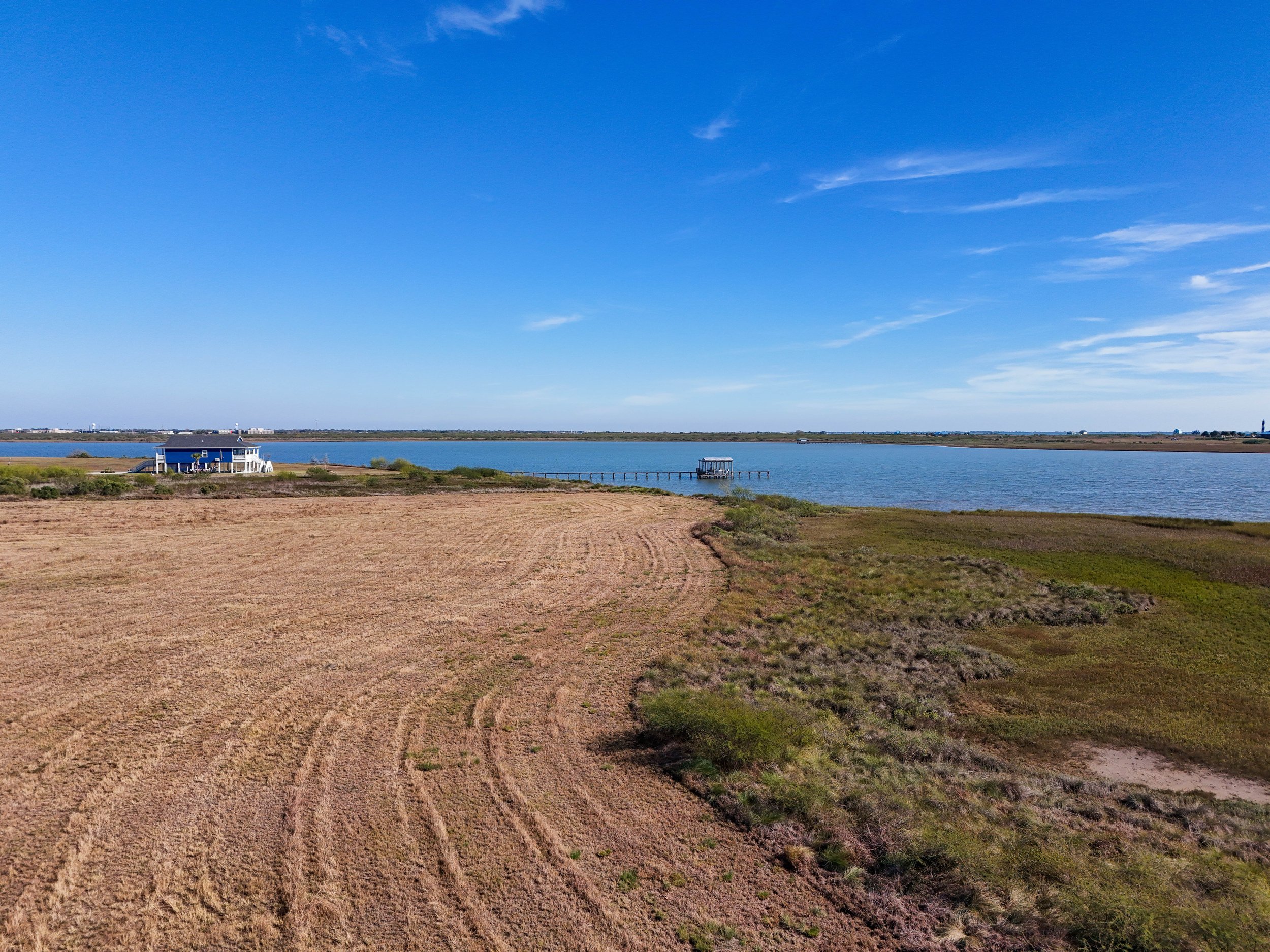 View of a large body of water with a house and a small dock on the shoreline, surrounded by open land and fields, under a partly cloudy blue sky.