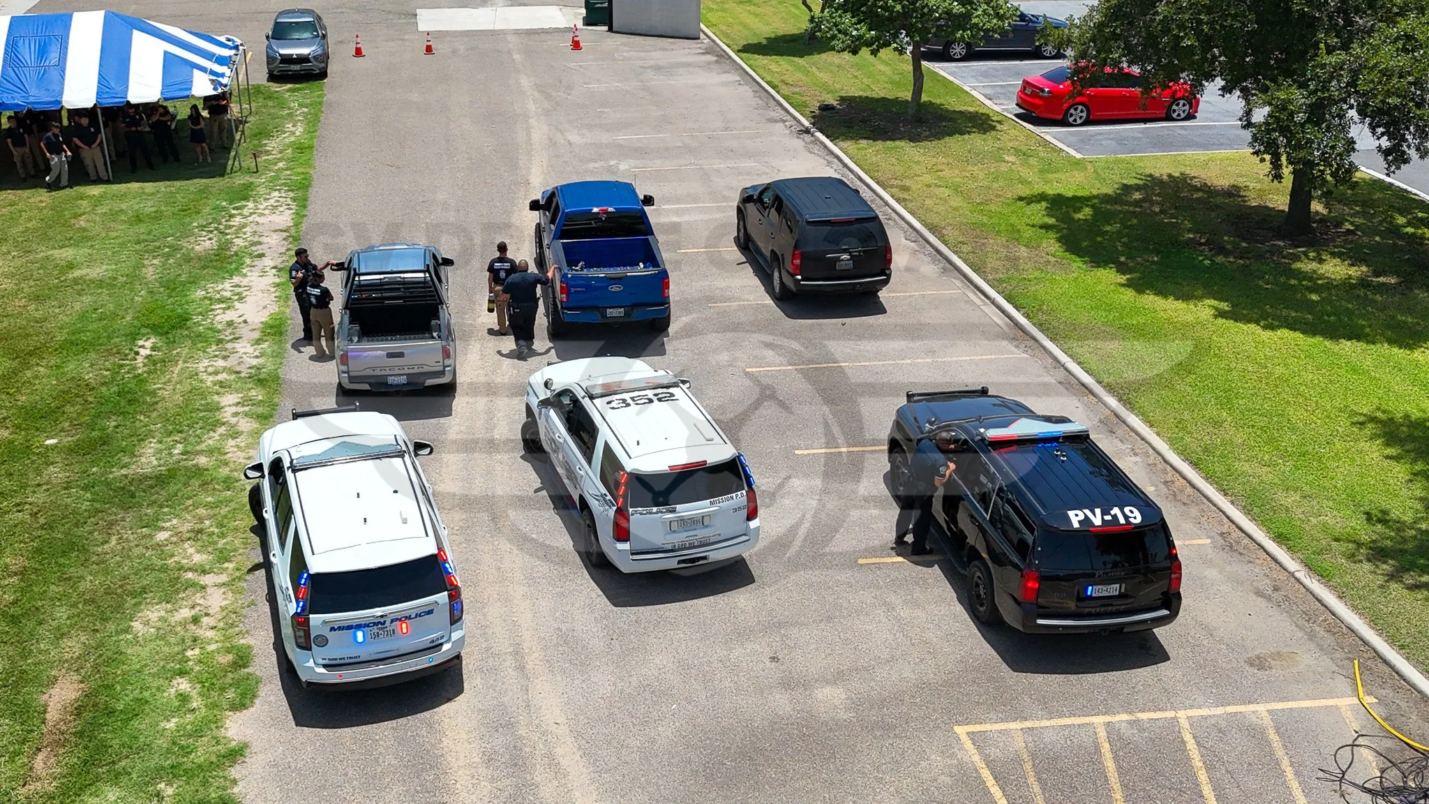 Several police vehicles parked in a lot with officers speaking to each other. Some officers are near the vehicles, and others are standing nearby. There is a tent on the left side with people inside, and a row of parked cars on the right side. The sc