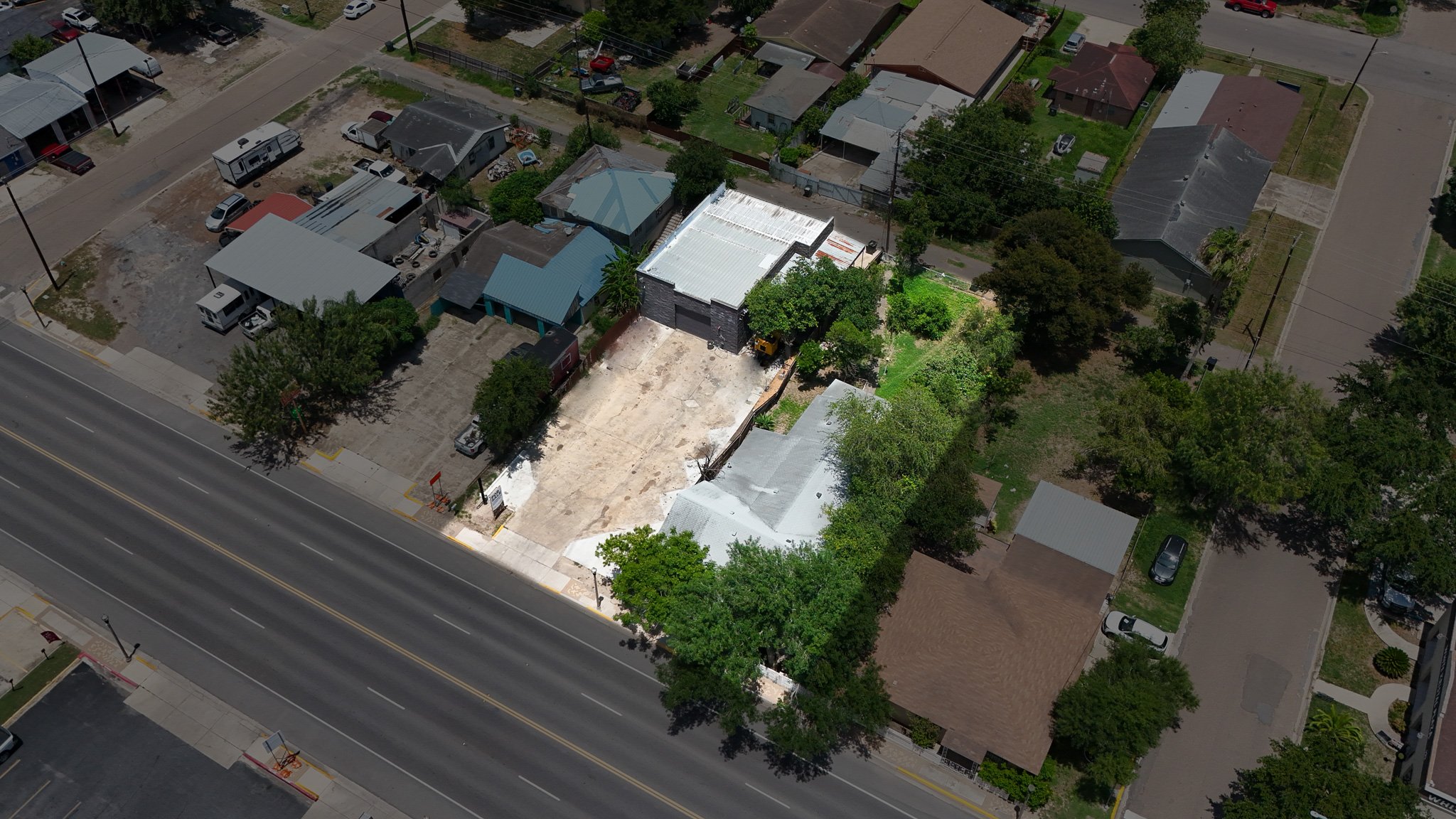 Aerial view of a residential neighborhood showing a commercial building with a white roof and a parking lot, surrounded by trees, houses with various roof colors, and streets with parked cars.
