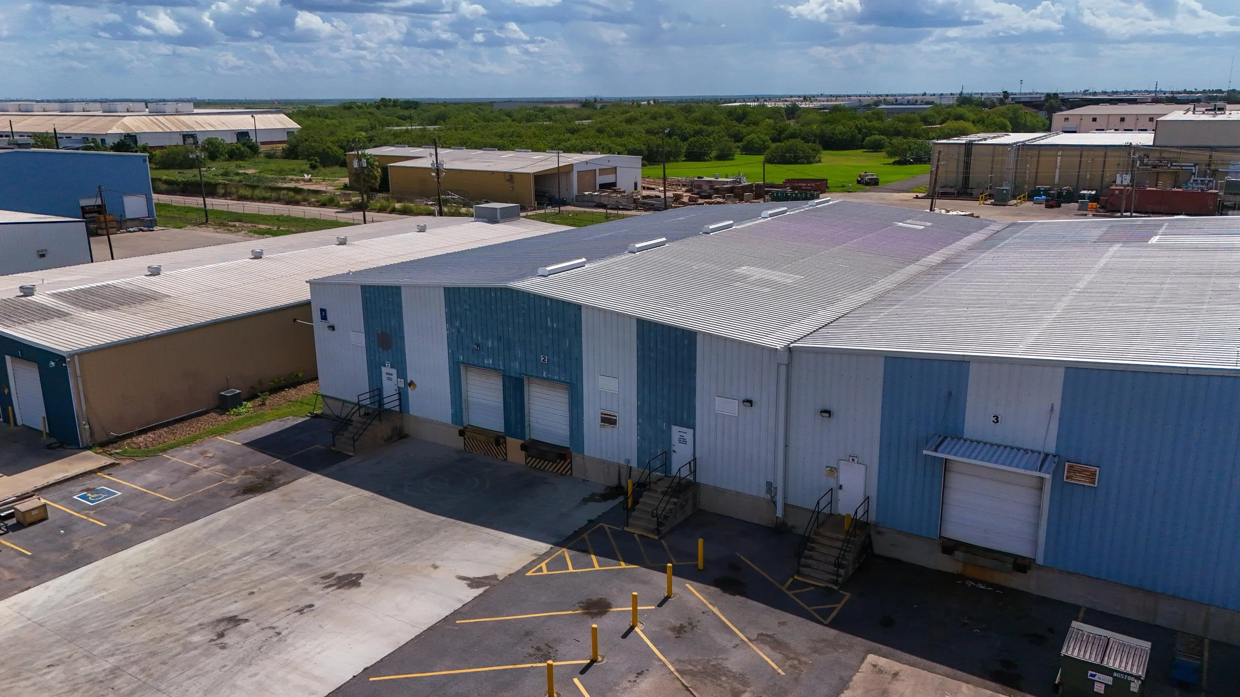 An aerial view of industrial warehouses with parking lots, loading docks, and surrounding greenery.