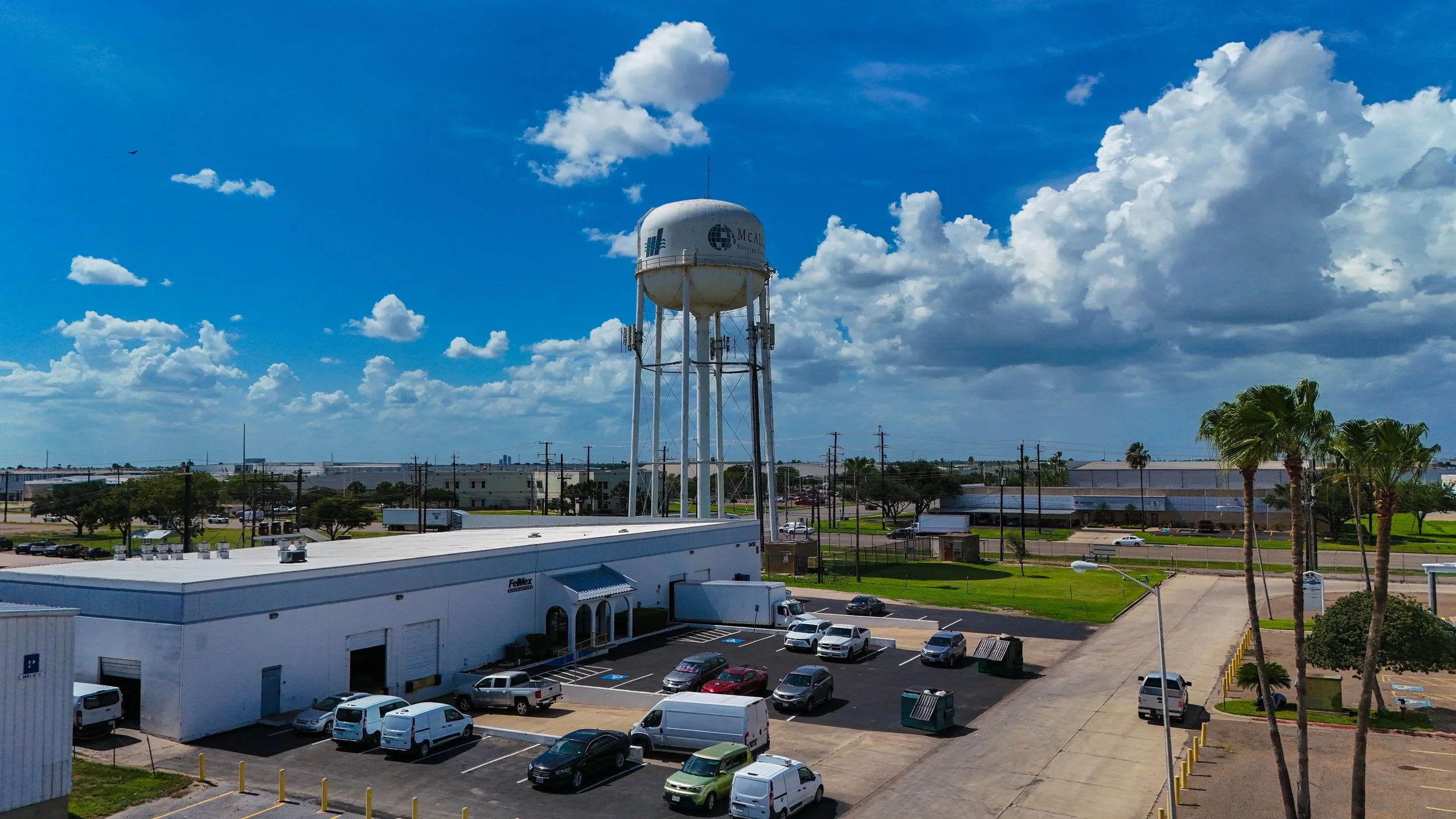 A water tower with the McAllen logo above a commercial building, parking lot with cars, and palm trees, under a partly cloudy blue sky.