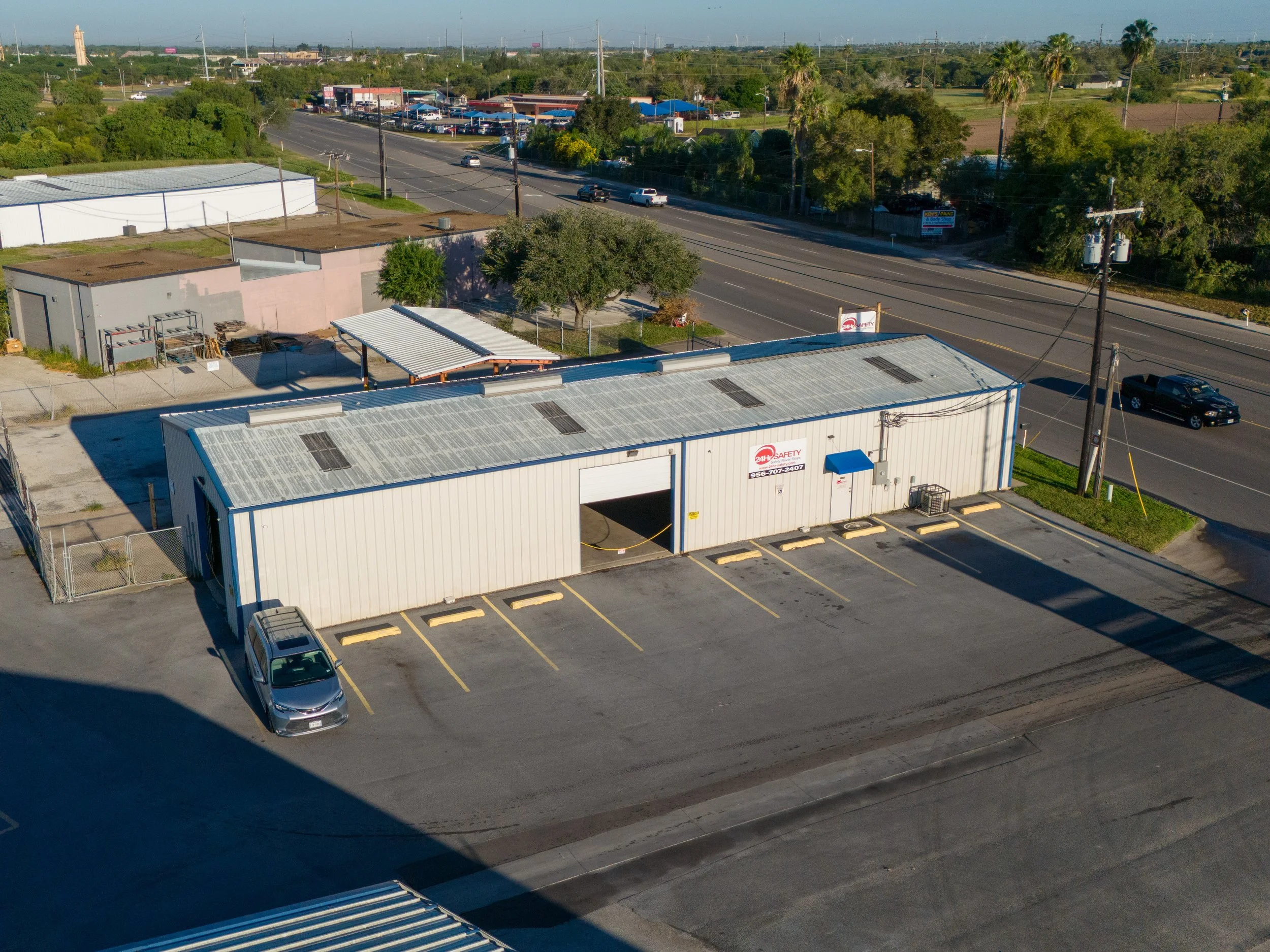 An aerial view of a commercial building with a parking lot, cars, and a road with moving vehicles, surrounded by trees and other buildings.