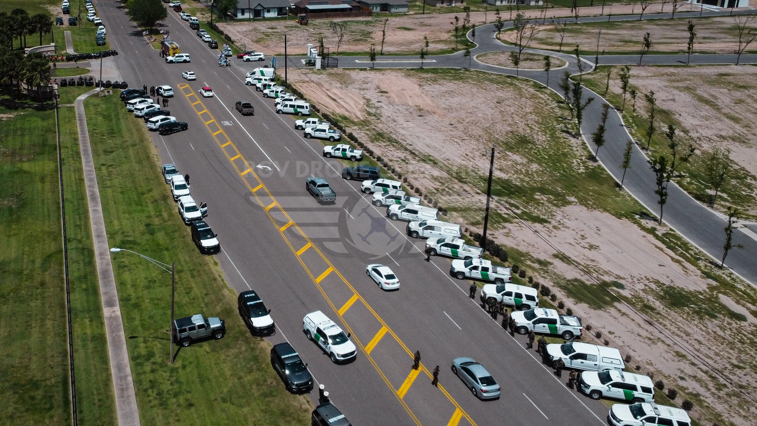 Aerial view of a police checkpoint with multiple police vehicles and officers on the road, with surrounding areas of grass, trees, and empty patches of land.