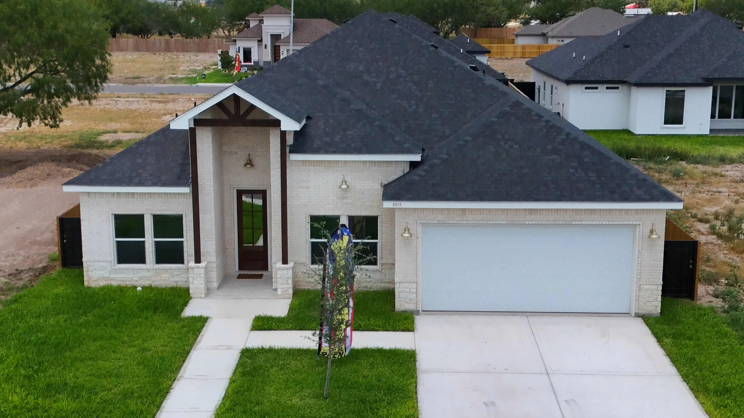 Newly built single-family house with white brick exterior, dark shingle roof, front porch, and attached two-car garage. Green lawn in front of the house with a sidewalk leading to the front door.