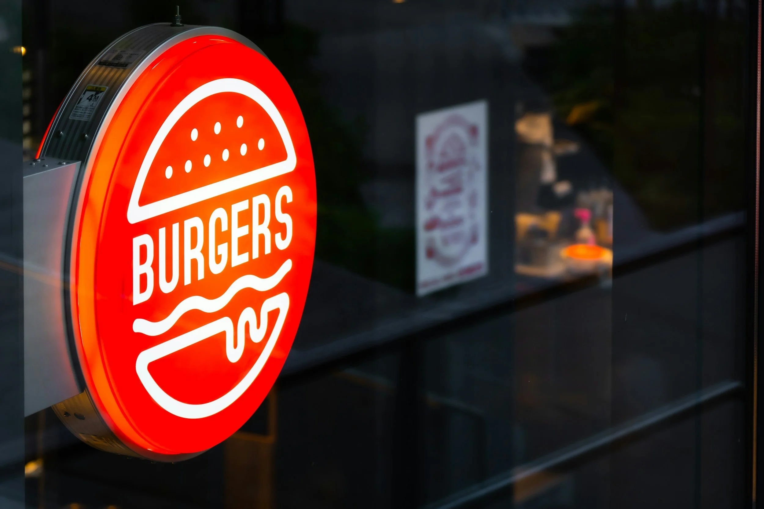Un panneau lumineux en forme de cercle avec un hamburger stylisé et le mot 'BURGERS' écrit en blanc, situé à l'extérieur d'un restaurant ou d'un fast-food.
