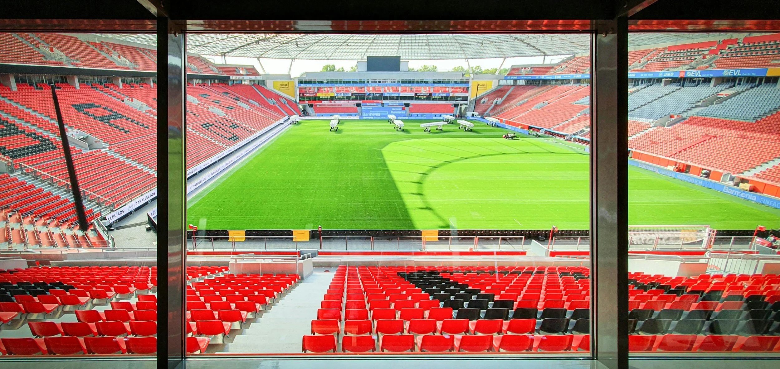 Vue intérieure d'un stade vide avec des sièges rouges, vue sur le terrain d'football vert et une grande enceinte, prise depuis une loge ou un balcon.