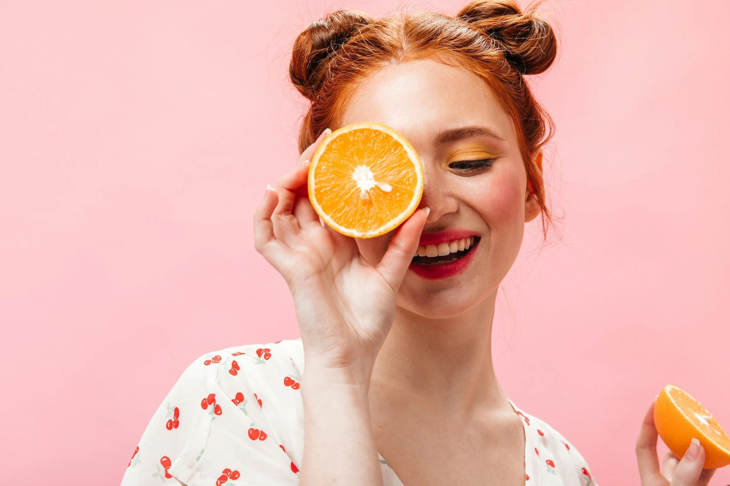 Jeune femme rieuse tenant une moitié d'orange devant son œil gauche, fond rose, vêtue d'une robe blanche à motifs de cerises rouges.