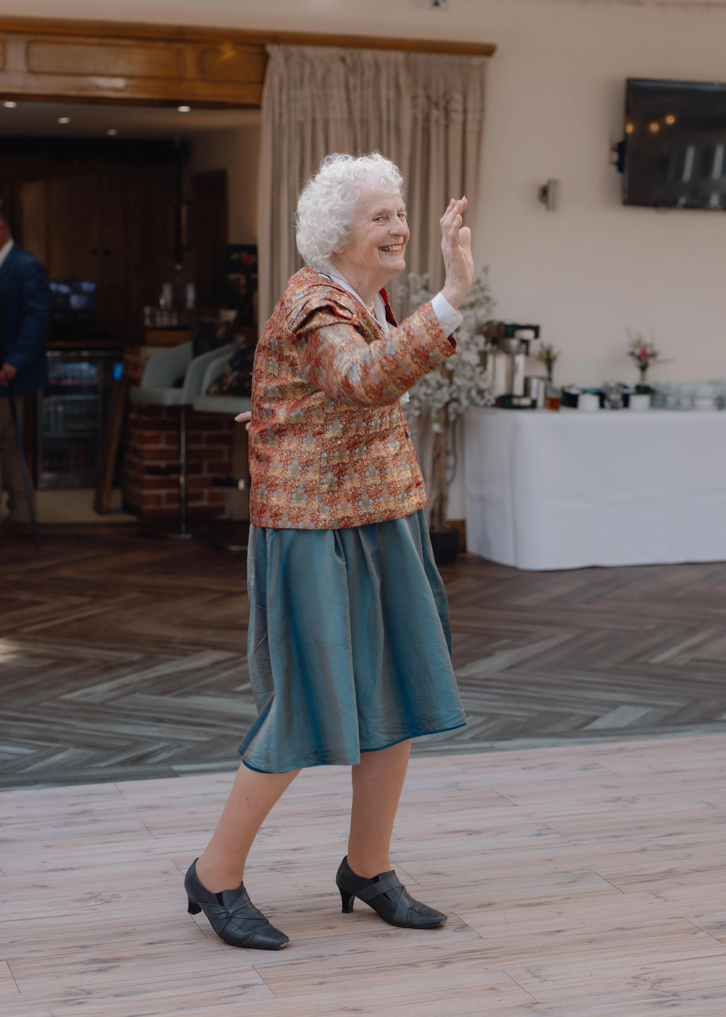 An elderly woman with curly gray hair dancing, smiling, and waving her hand in a room with wooden floors, beige curtains, and a white table with decorations in the background.