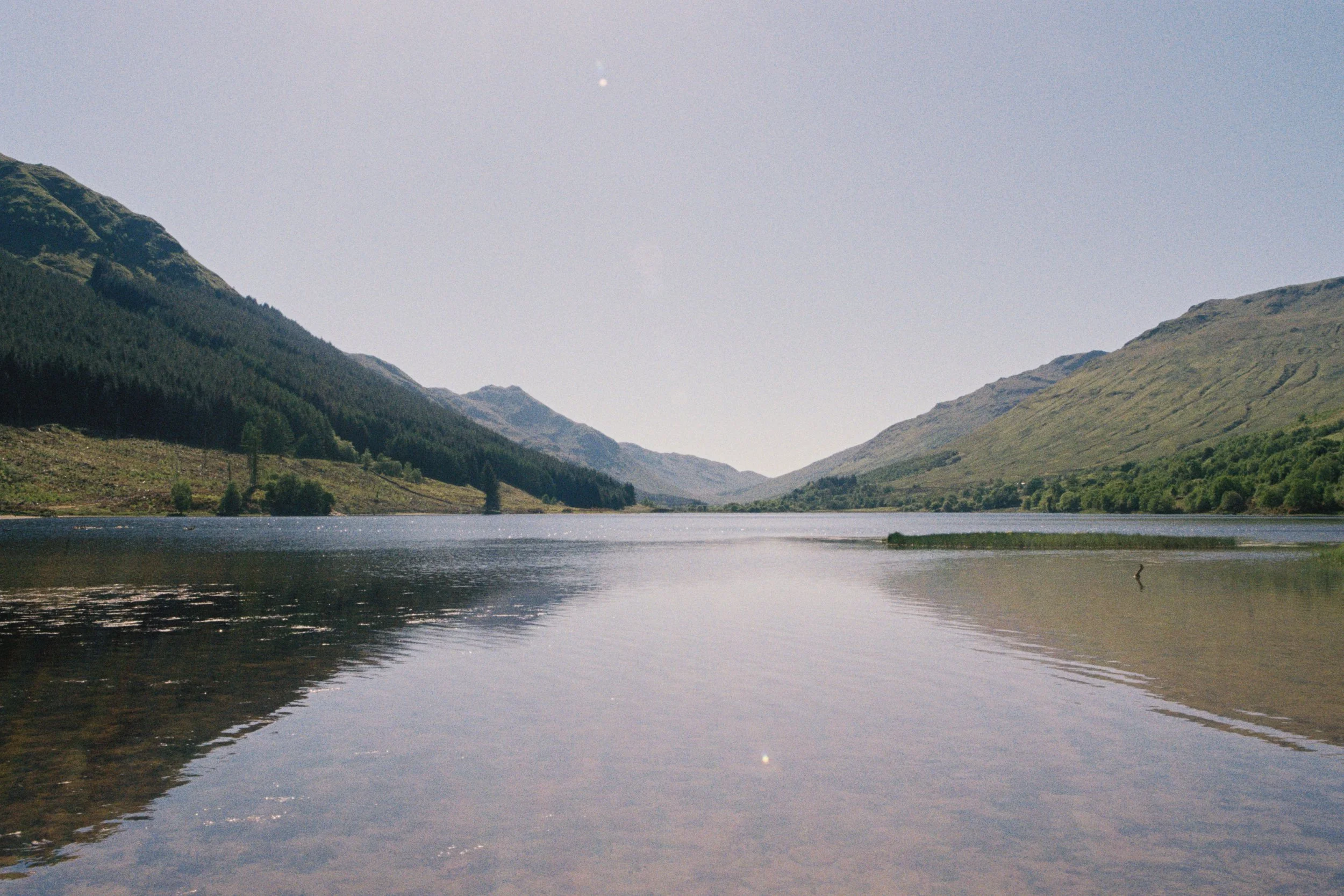 Calm river reflecting green rolling hills and mountains under a clear blue sky with no visible clouds.