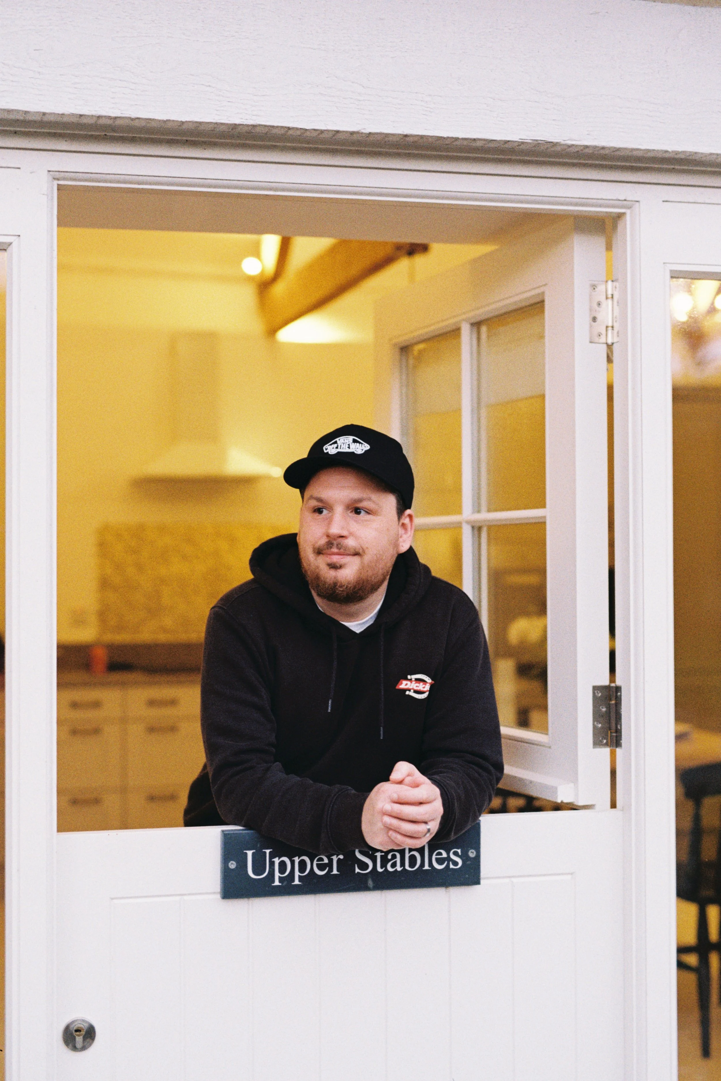 A young man with a beard and mustache, wearing a black hoodie and a black baseball cap with a white logo, is standing behind a white window frame that has a sign reading "Upper Stables." He is leaning on the windowsill with his hands clasped, looking