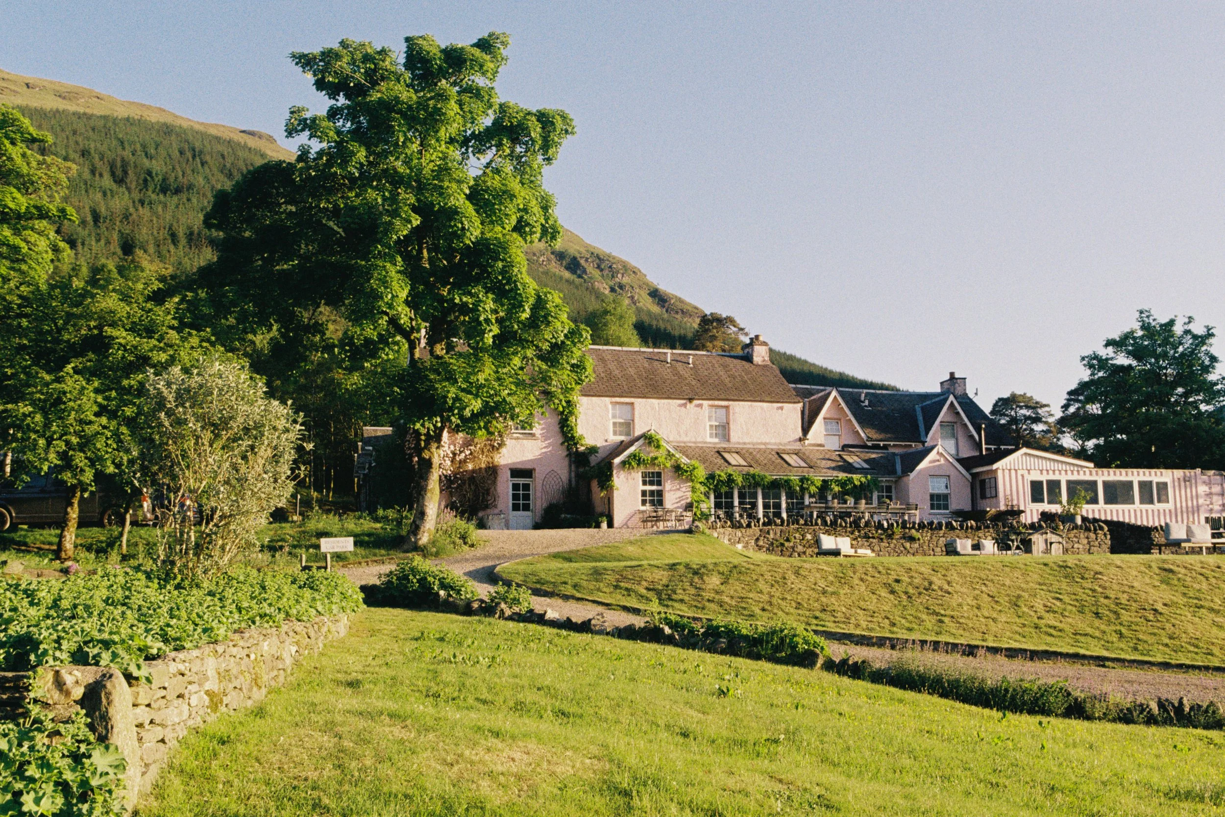 A scenic view of a garden with neatly maintained lawns, shrubs, and flowers, leading to a light pink house with a patio. Behind the house, there are houses and a hillside covered with lush green trees and a mountain in the background, under a clear b