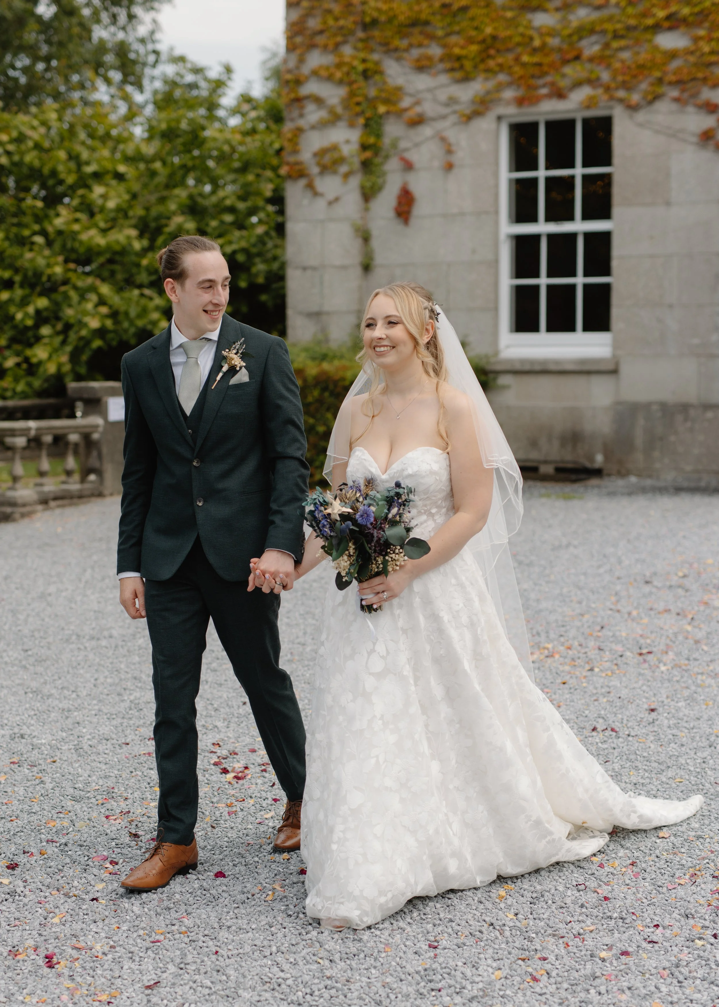 A happy bride and groom holding hands smiling outdoors near a stone building with leafless and green trees in the background.