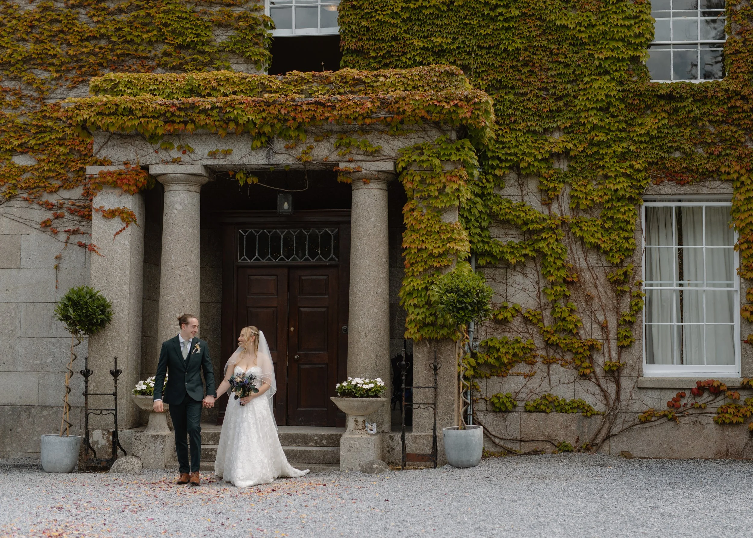 A bride and groom holding hands and smiling outside a historic building with ivy-covered walls, large potted plants, and a wooden door.